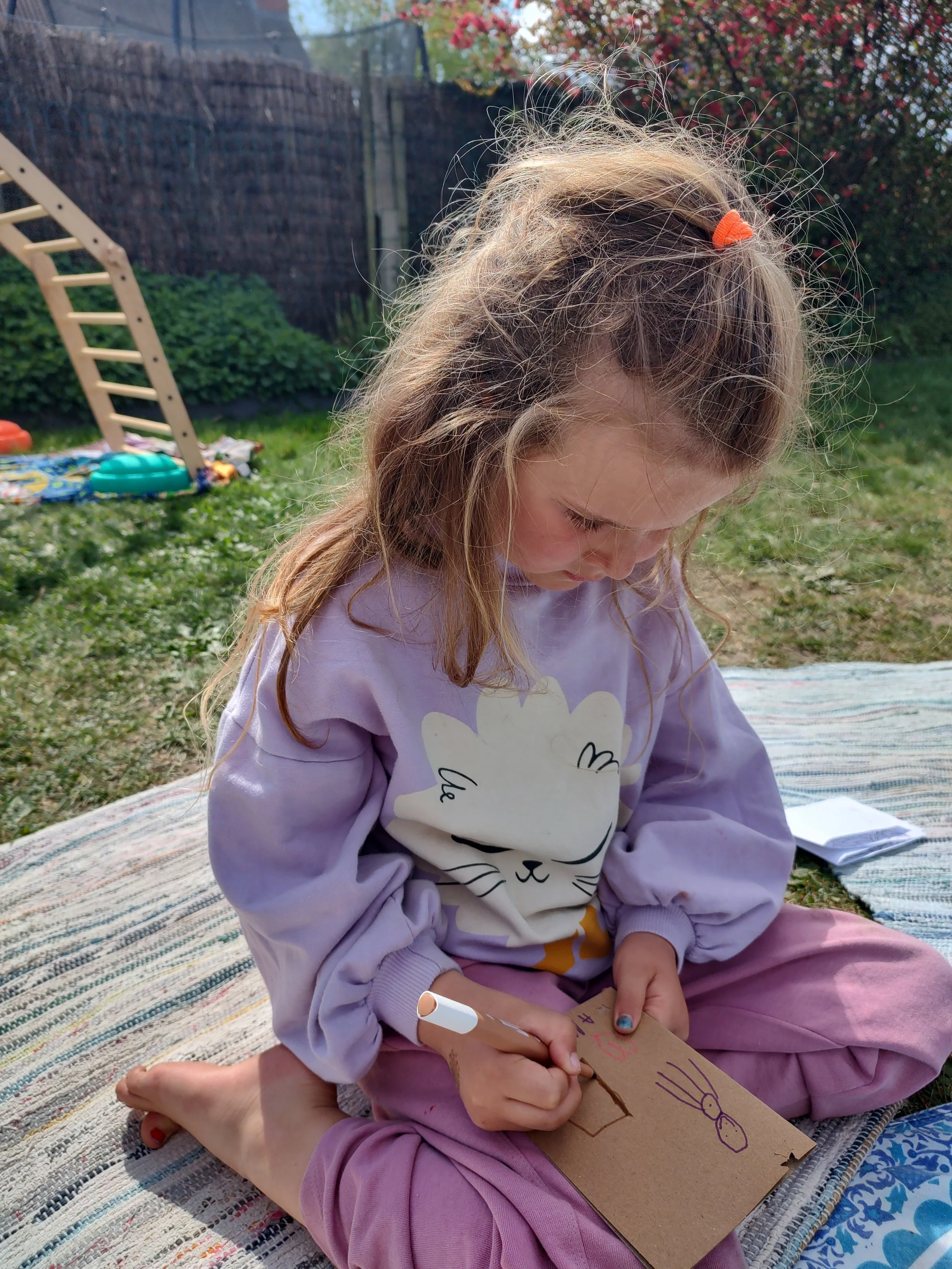 A young girl with messy blonde hair wearing a purple sweatshirt with a cat face design, pink pants, and a small orange hair tie, sits cross-legged on a woven blanket in a garden, drawing on a piece of cardboard with markers. She is focused on her art