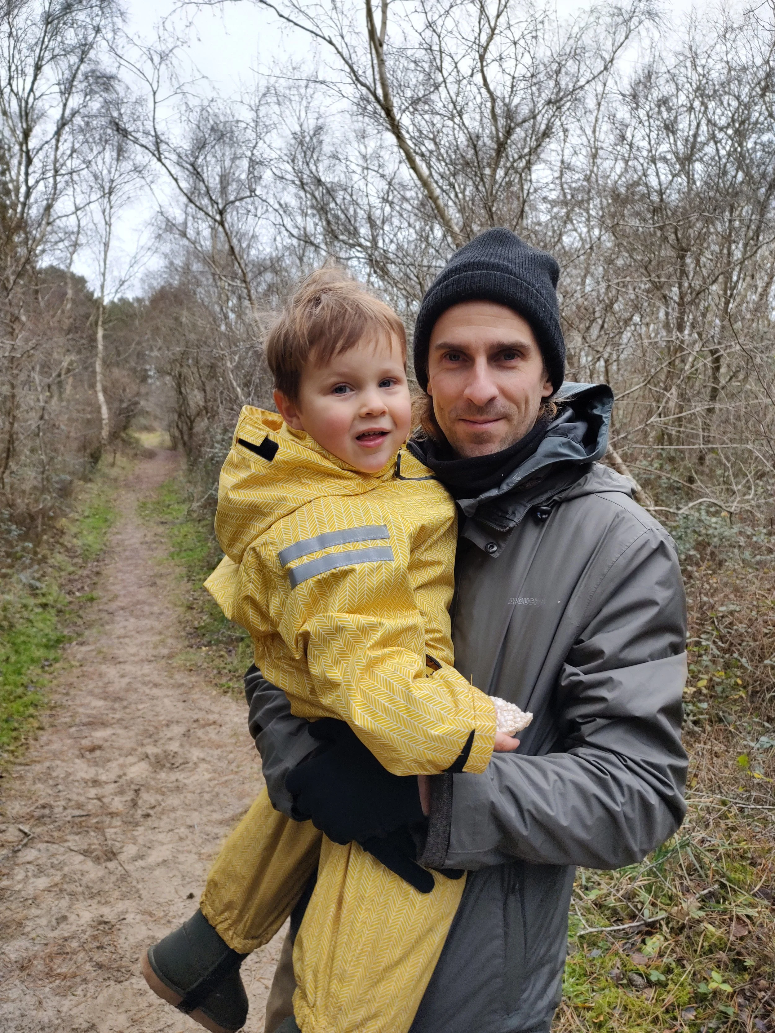 A man in a gray jacket and black beanie holds a young boy in a yellow jacket on a dirt trail surrounded by leafless trees.