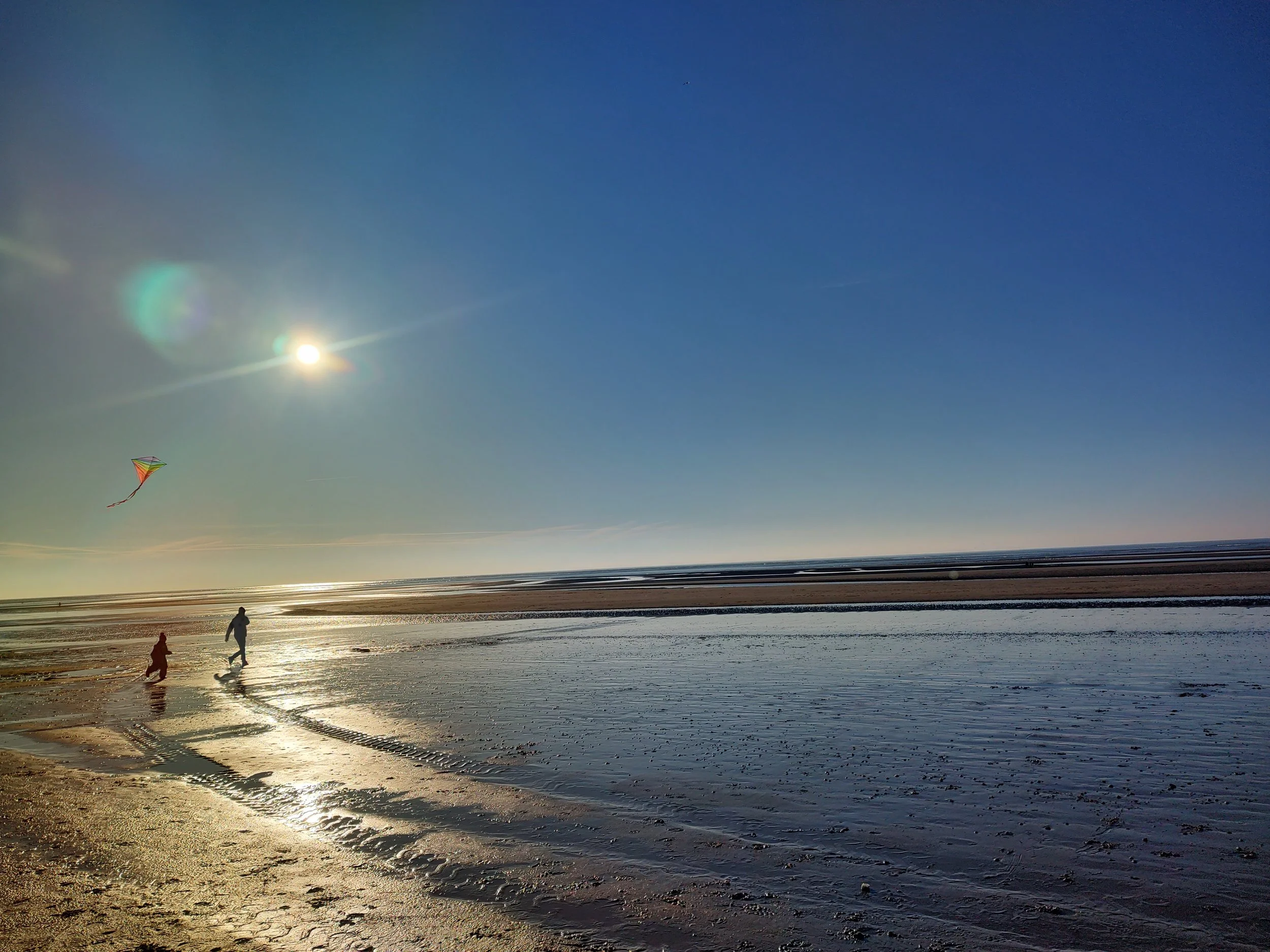 People flying a kite on a beach during sunset with calm waters, a clear blue sky, and the moon visible in the sky.