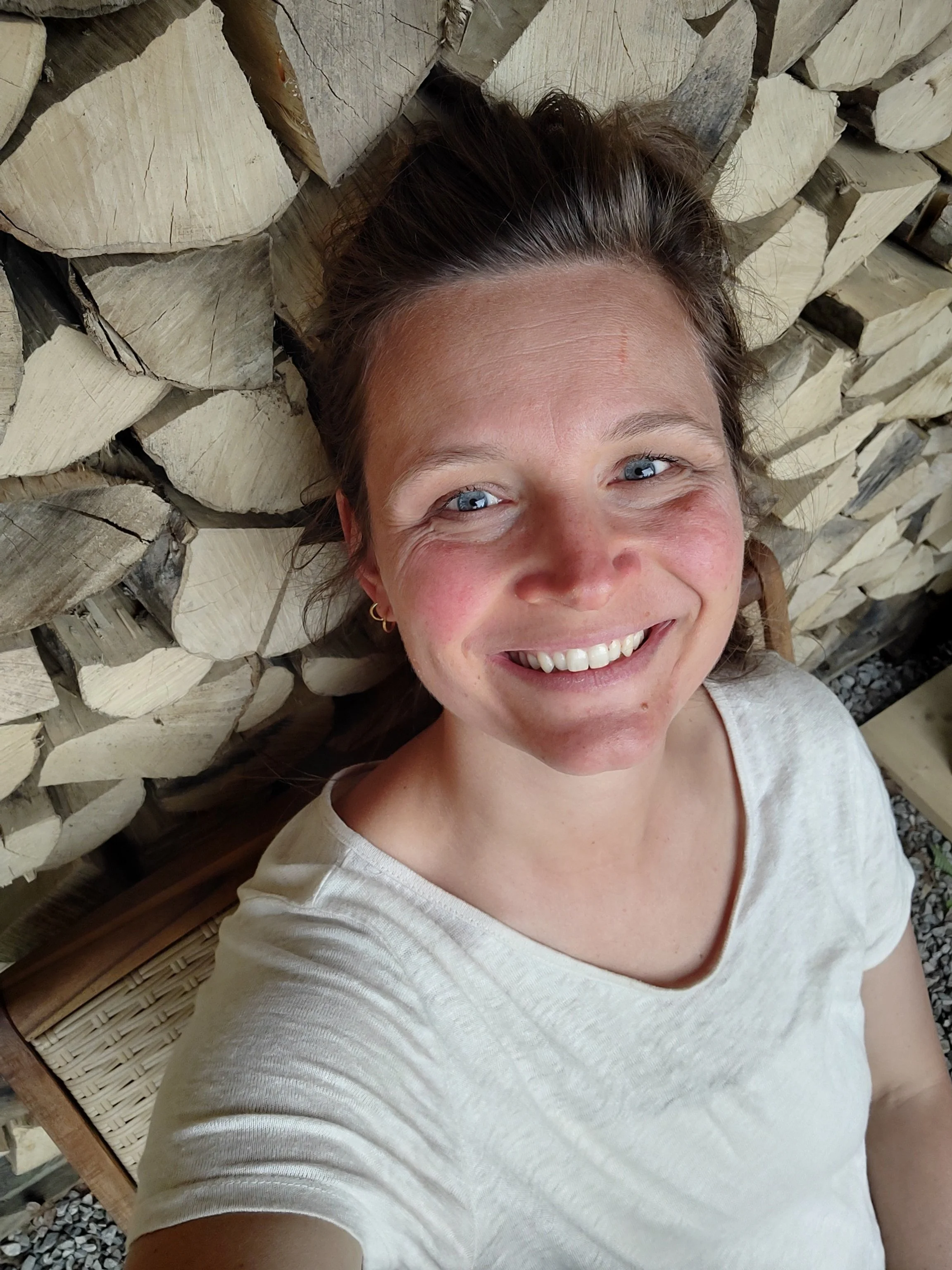 A woman with light skin, blue eyes, and brown hair lying on a stack of cut wood logs, smiling at the camera.