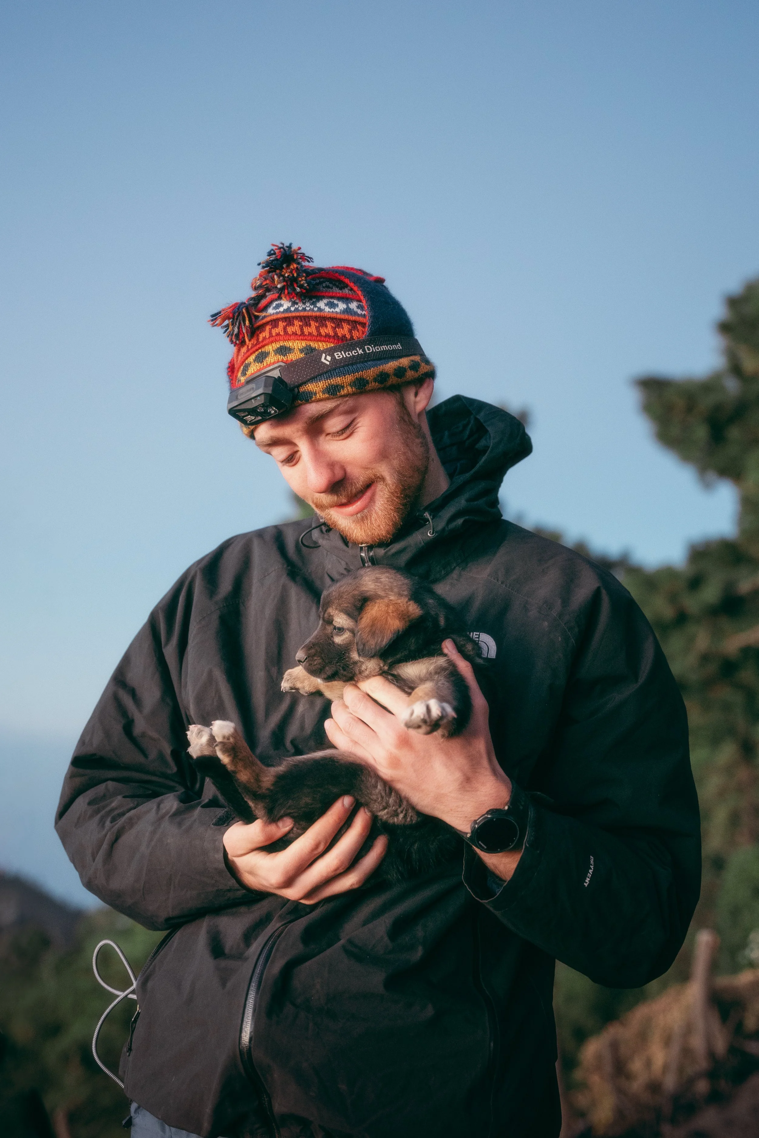A man in outdoor clothing holding a small puppy, with a landscape and trees in the background.