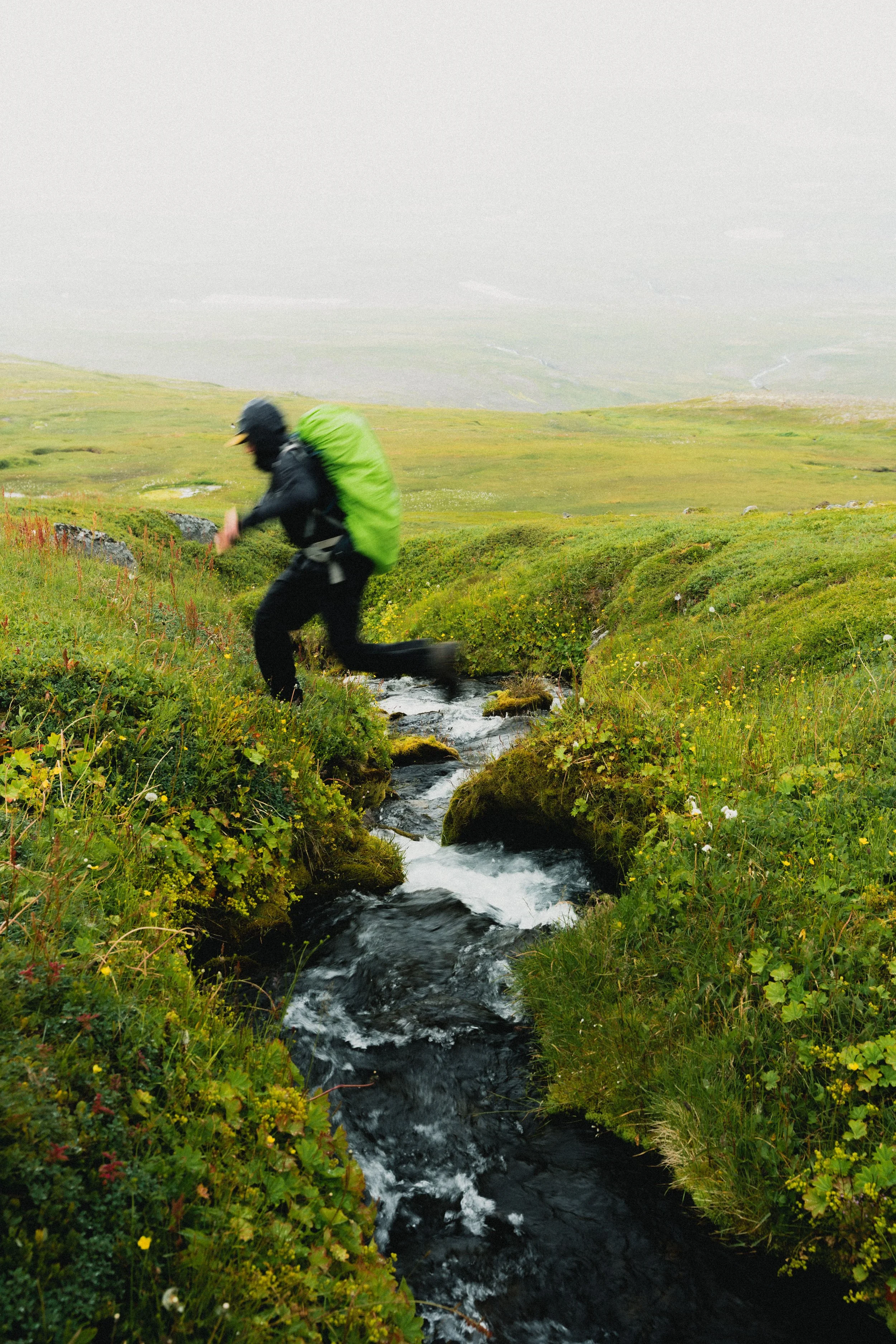 A person with a green backpack and a black rain jacket is crossing a small stream in a lush, green meadow under a cloudy sky.