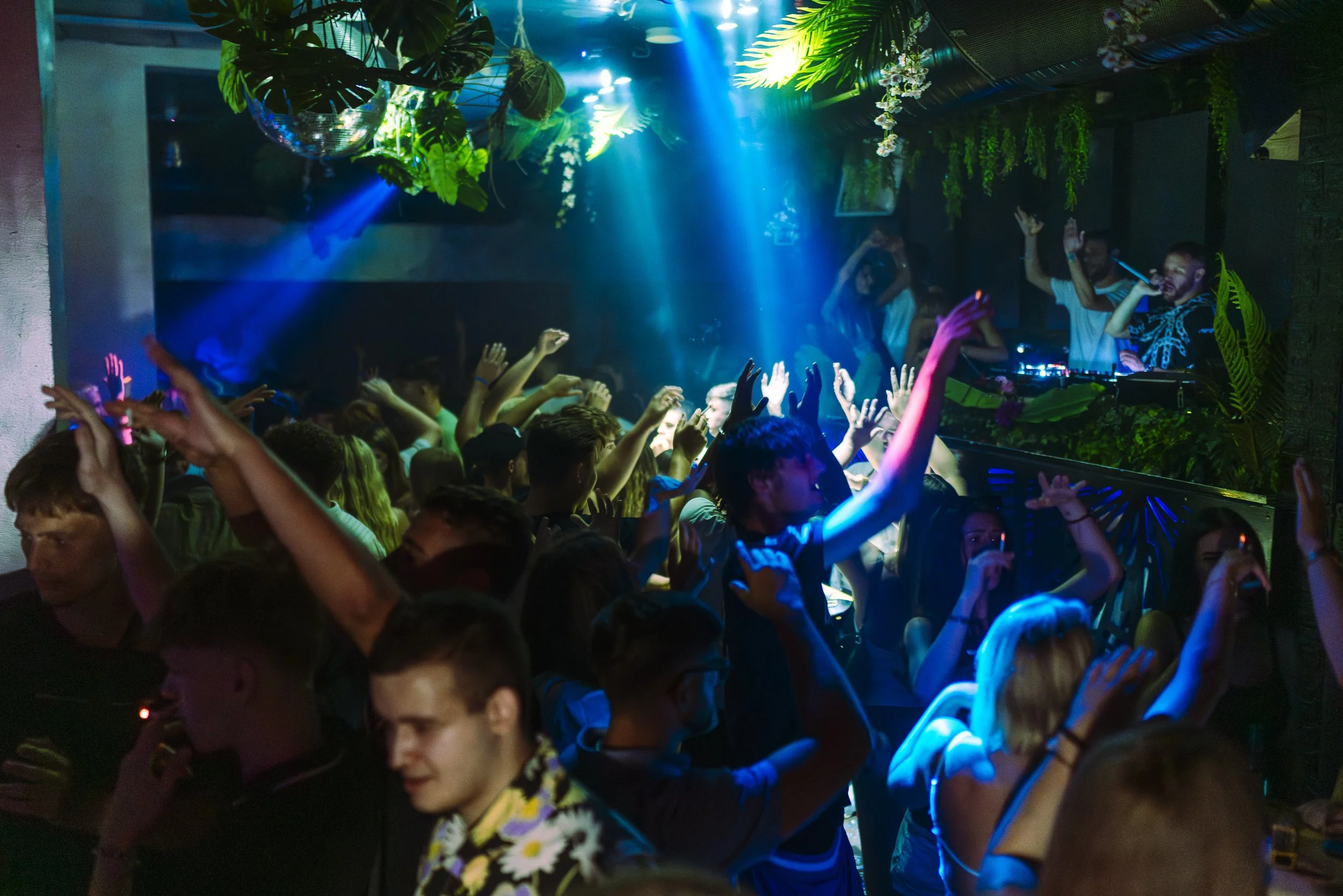 Crowd of people dancing and raising their hands in a nightclub with a DJ performing on stage, surrounded by colorful lights and tropical plants.