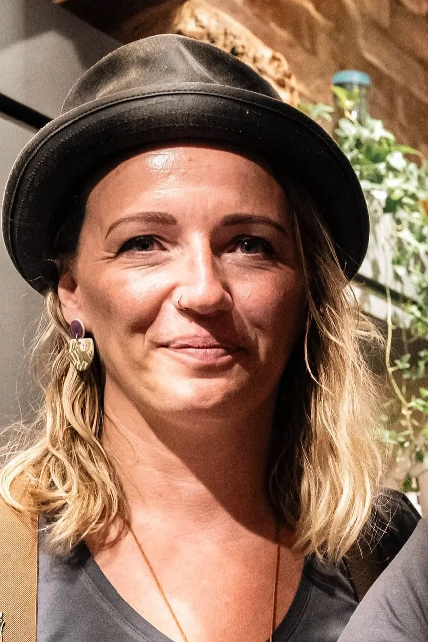 Eine Frau mit blonden, lockigen Haaren trägt einen schwarzen Hut und große Ohrringe, im Hintergrund sind Pflanzen und eine Ziegelmauer sichtbar.