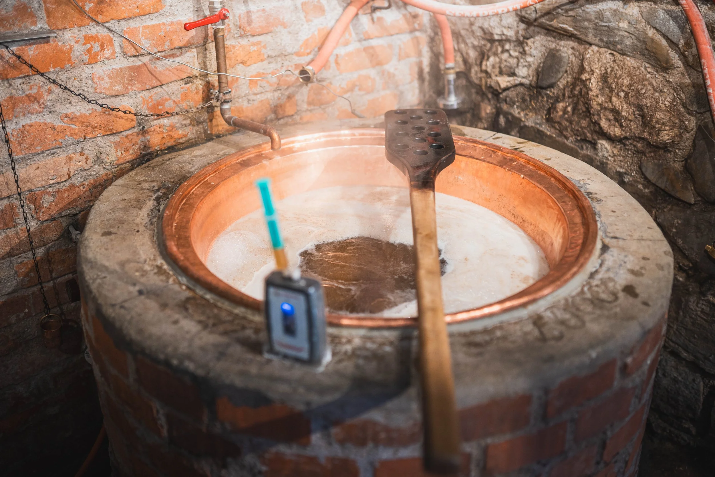 Ein Kupferkessel auf Steinen, in dem eine heiße Flüssigkeit schäumt, mit einem Holzschlägel und einem Metallrost darauf. Im Hintergrund sind Wasserleitungen und eine Wand aus Ziegelsteinen.