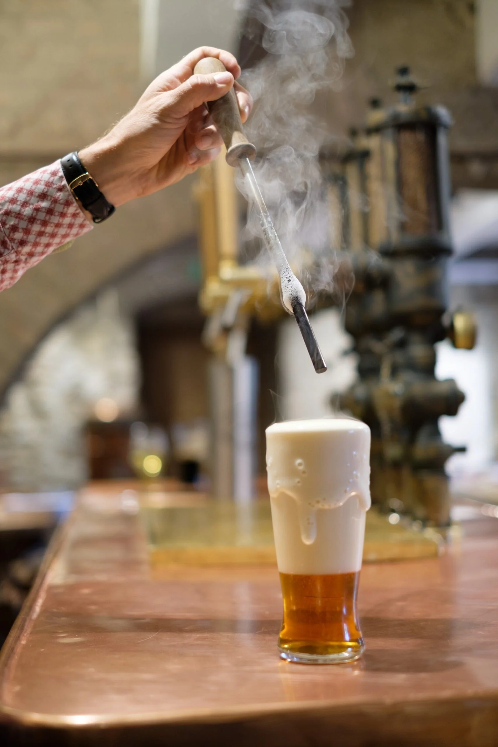 Ein Bierglas, das mit Rauch bedeckt ist, während eine Person mit einem Rührstab eine Bunsenbrenner-artige Flamme über das Glas hält.