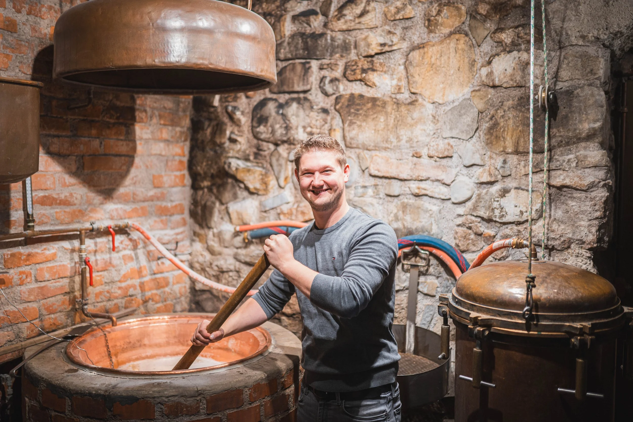 Ein lachender Mann beim Arbeiten in einer traditionellen Brennerei, die aus Stein und Backstein gebaut ist, mit Kupfer- und Metallgeräten.