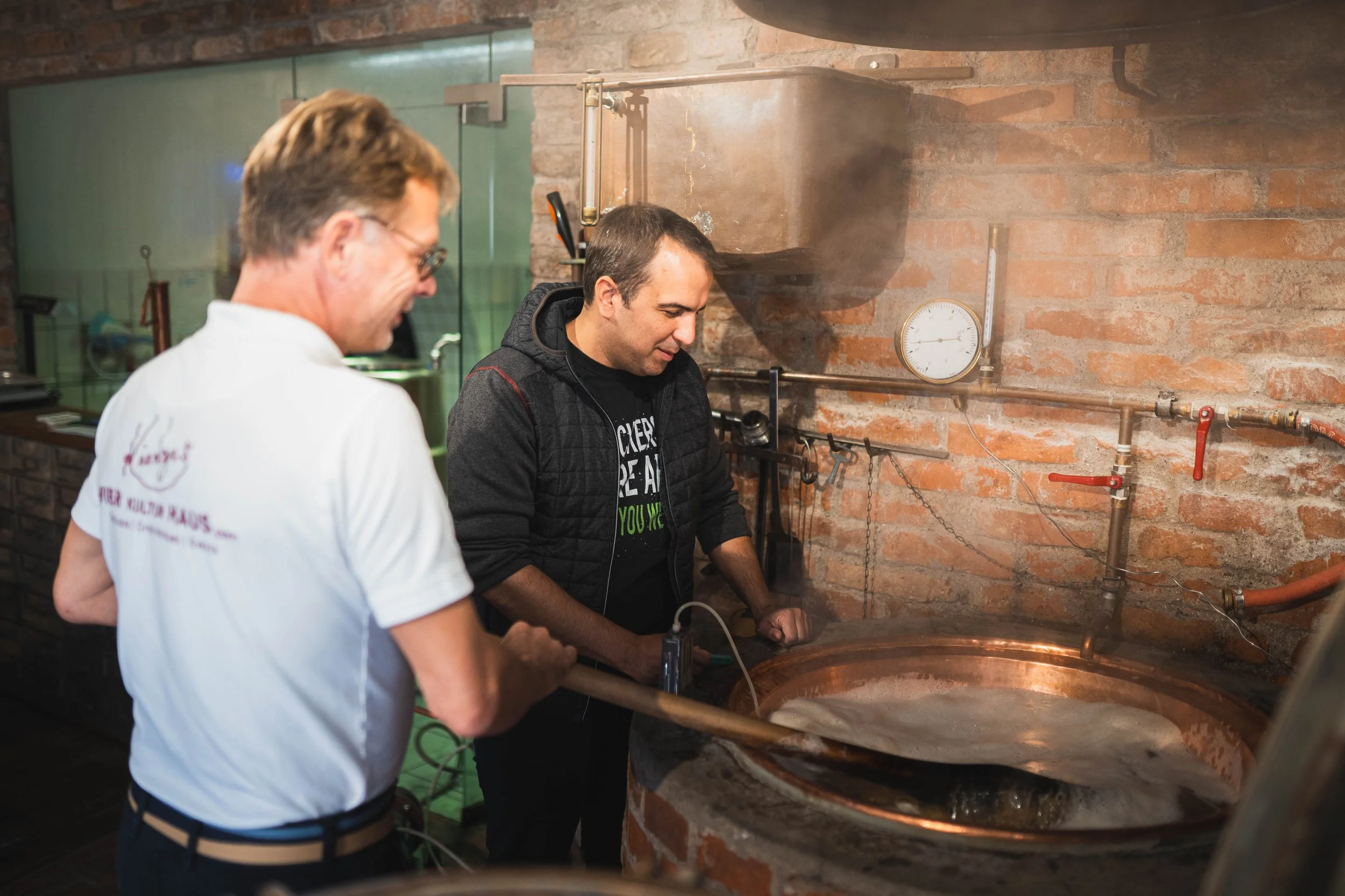 Zwei Männer beim Brauen in einer Brauerei, einer rührt in einem großen kupfernen Braukessel, der andere beobachtet.