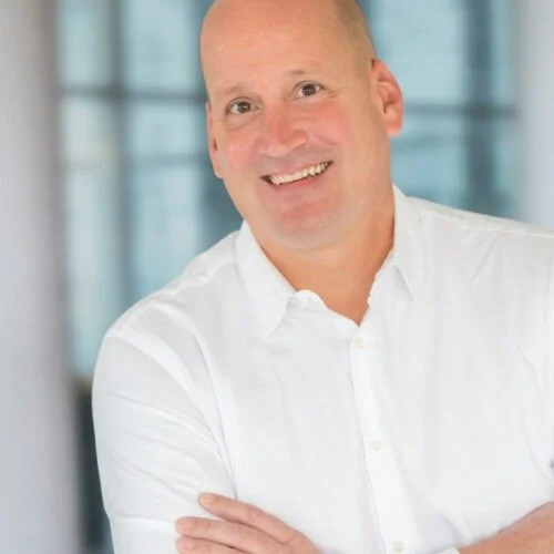 Ein lächelnder Mann mit kurzen Haaren in einem weißen Hemd vor einem modernen Bürogebäude.