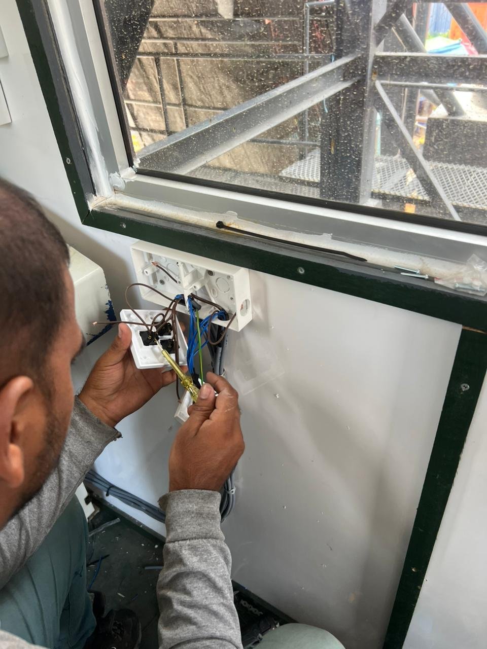 A man working on electrical wiring inside a room near a window. The wiring is connected to a white electrical box, and the window has rain droplets on it.