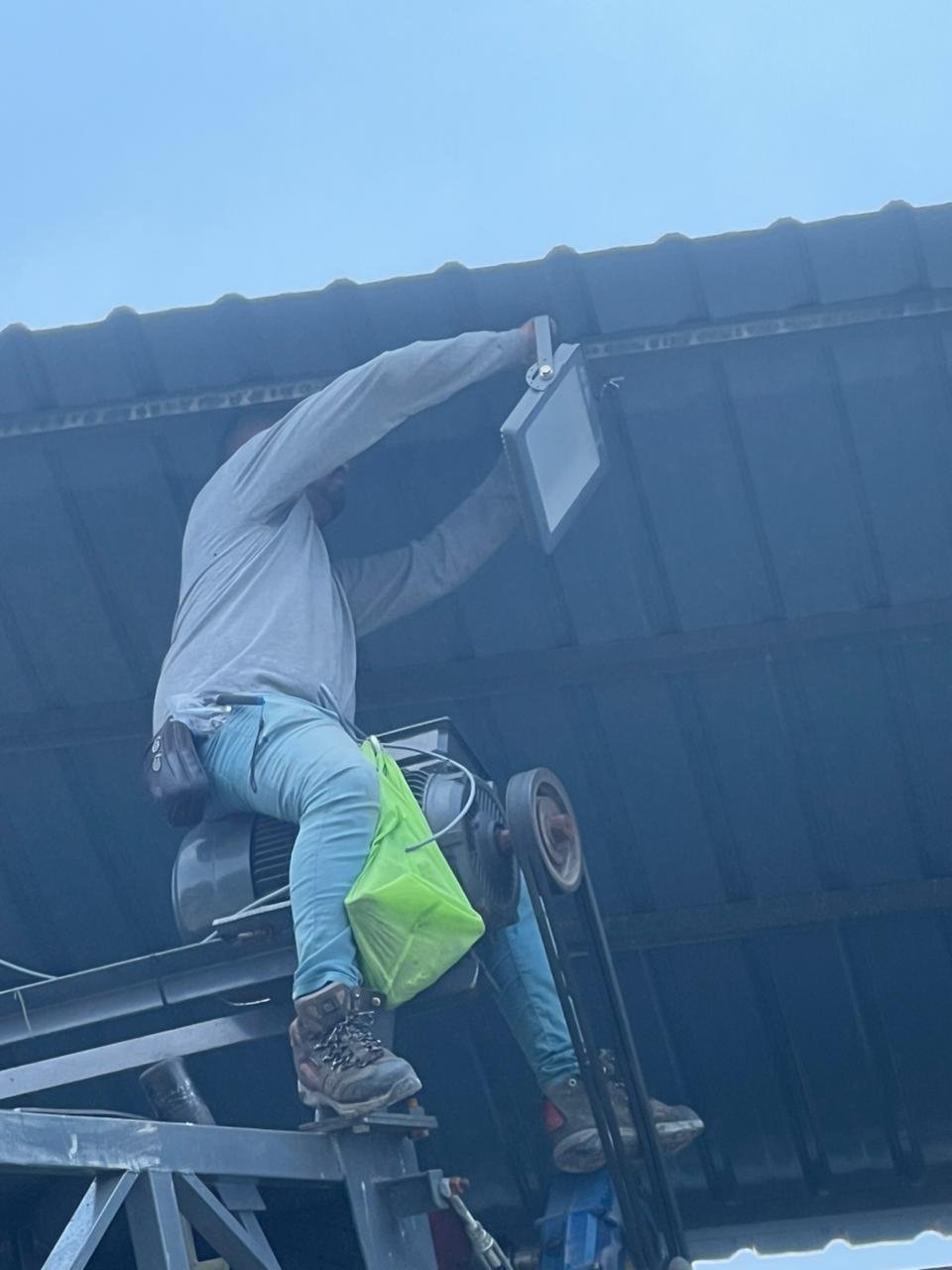 A worker is standing on a lift, installing or repairing an outdoor light fixture under the roof of a building.