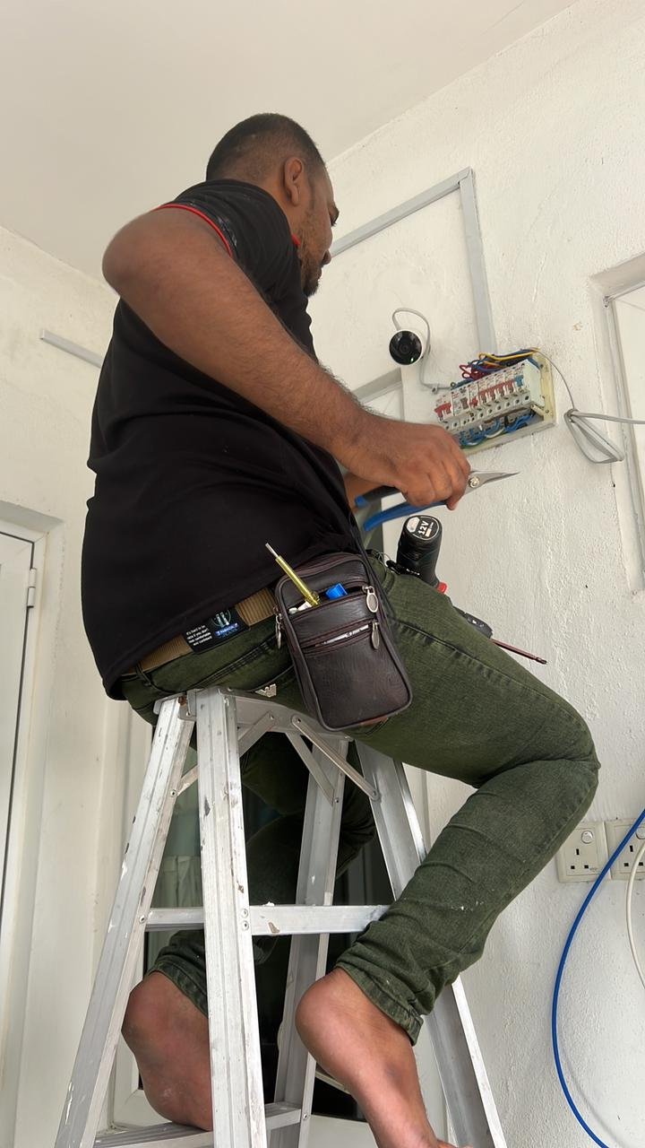 A man working on an electrical panel on a white wall, sitting on a step ladder. He is wearing a black shirt and green pants, with tools attached to his belt.