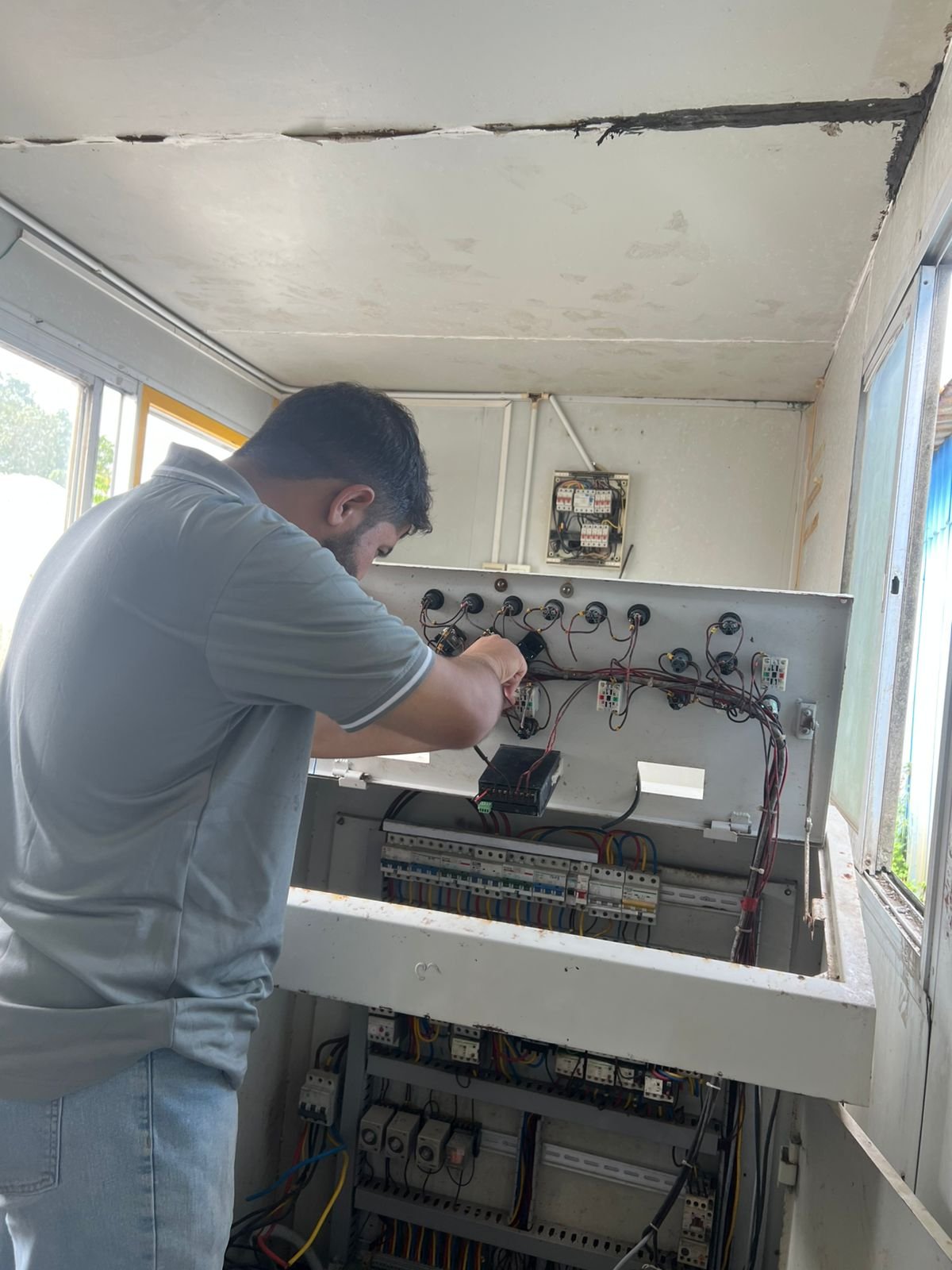 A man working on the wiring inside an electrical panel or circuit box in a room with windows.
