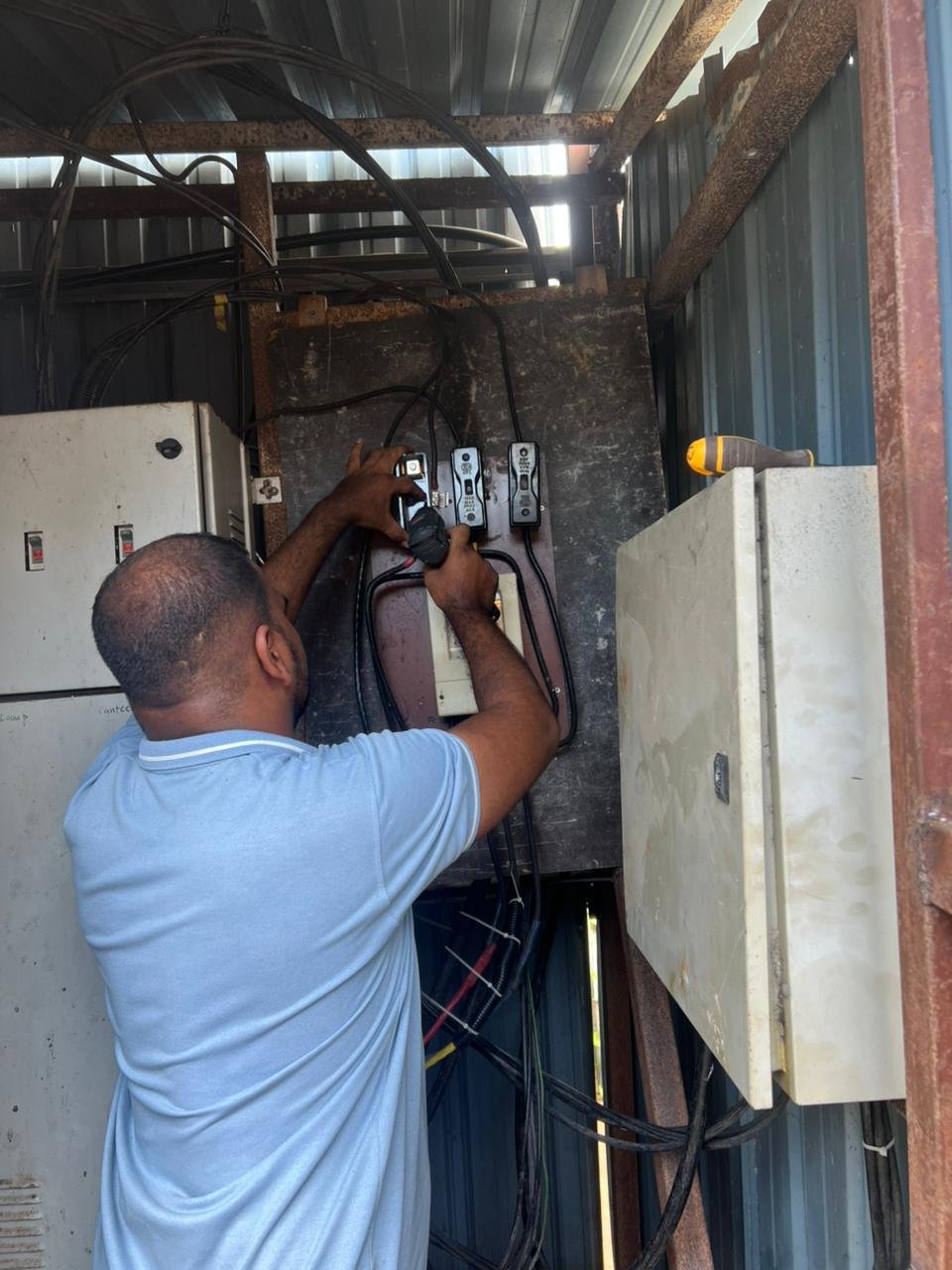 A man working on electrical wiring inside a metal shed. He is using a screwdriver or similar tool, with various electrical wires and switches visible around him.