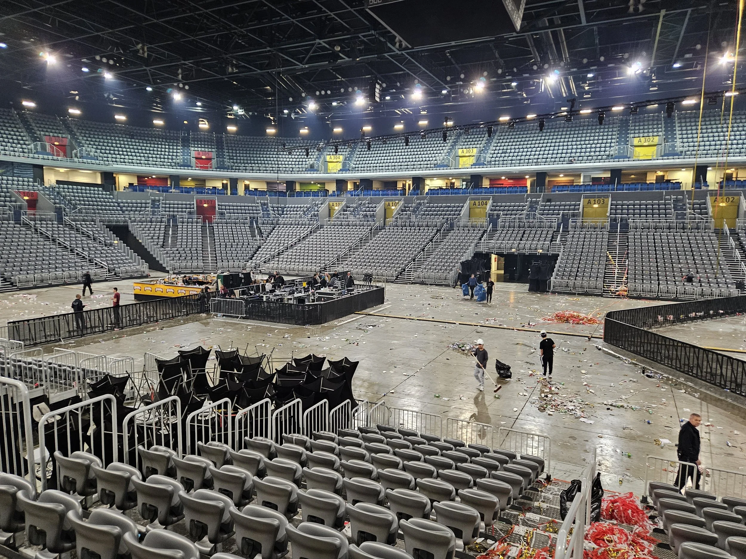 Empty indoor arena with scattered debris, stage equipment, and personnel cleaning up after event, with rows of empty seats and stadium lighting.