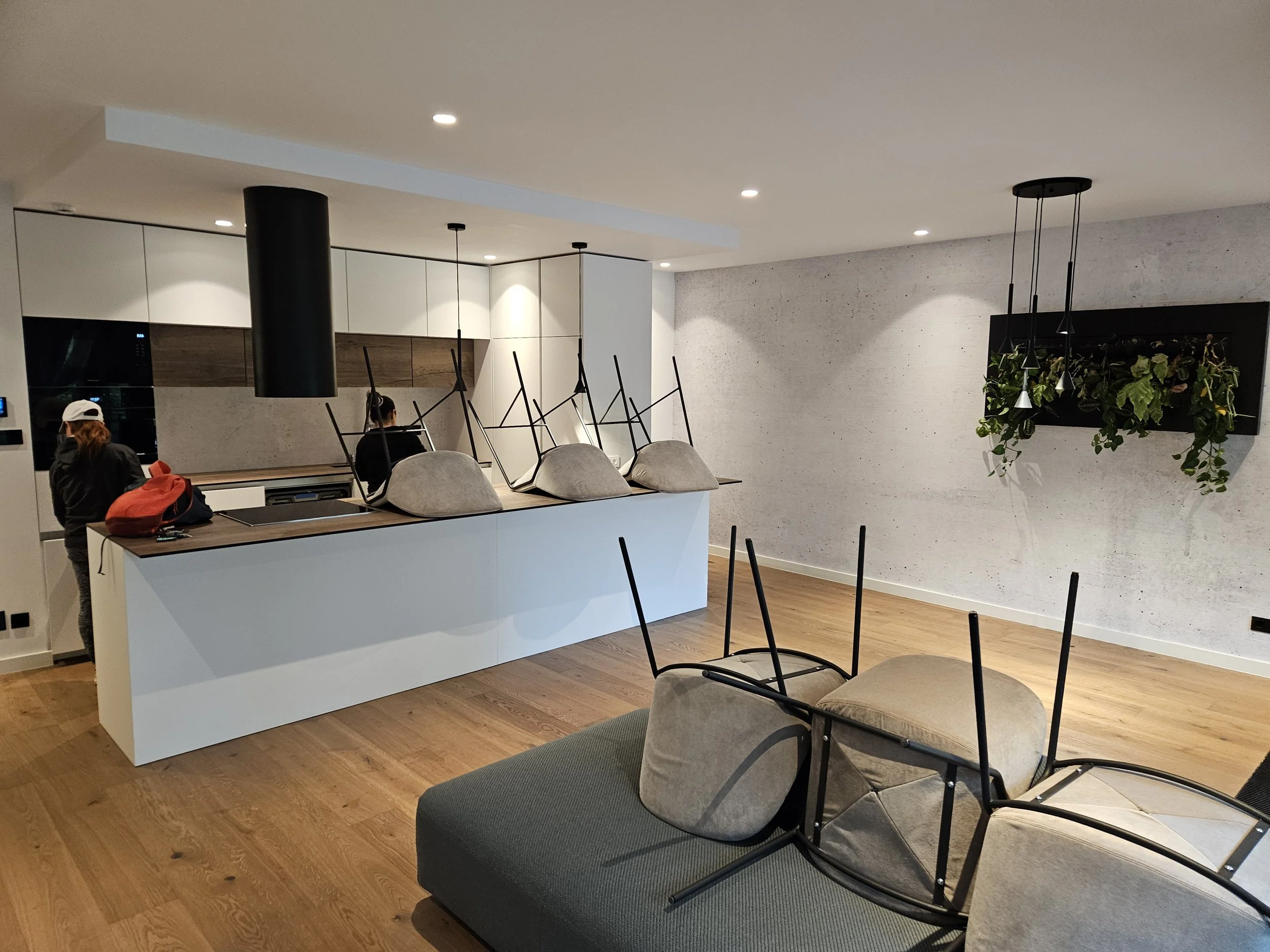 Modern kitchen and living room with chairs on top of the counter, a wall with a plant decoration, and seating area with chairs and a sofa.