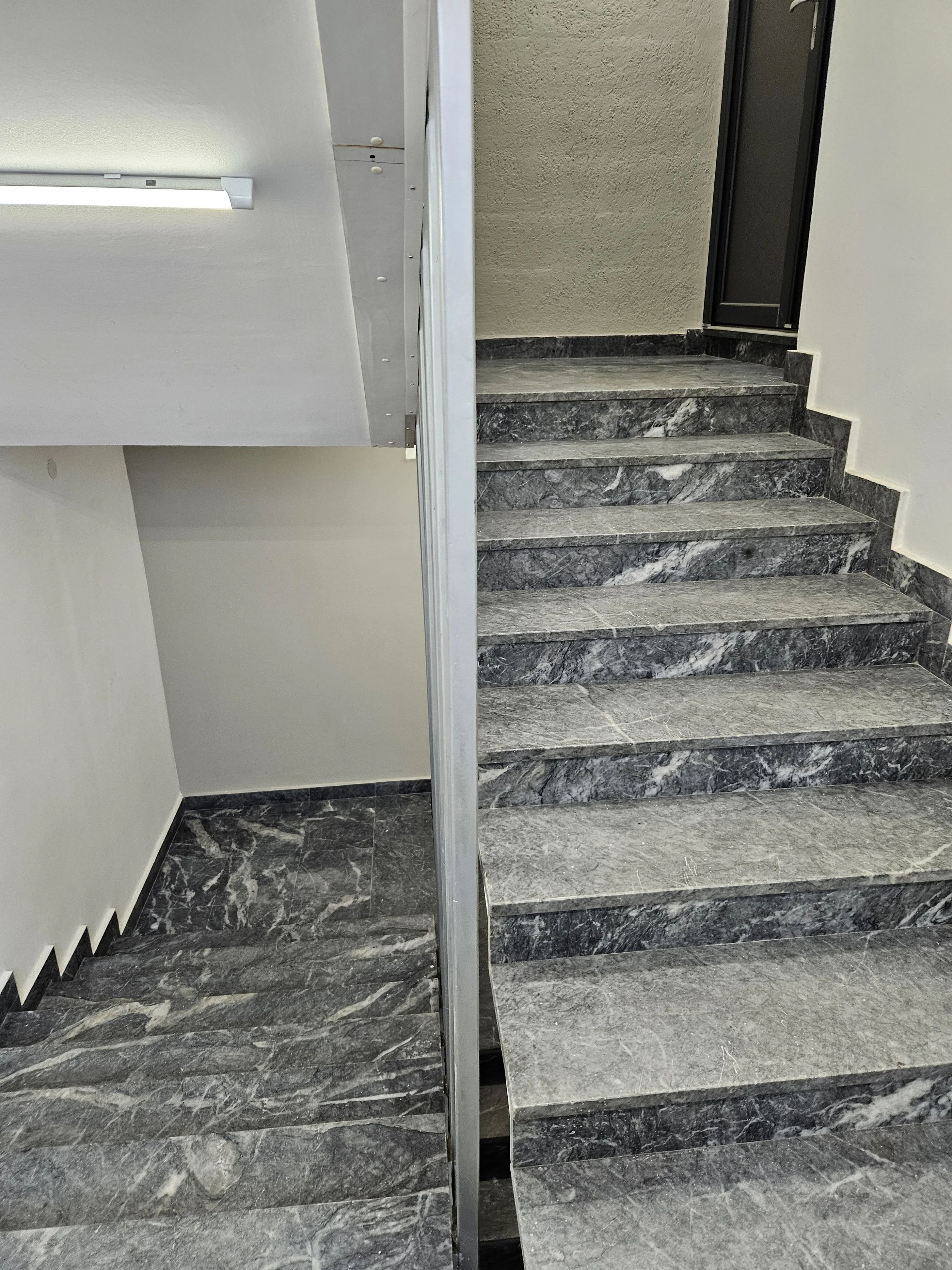 Indoor staircase with dark gray marble steps, metal railings, and a partial view of a white wall and the entrance door.