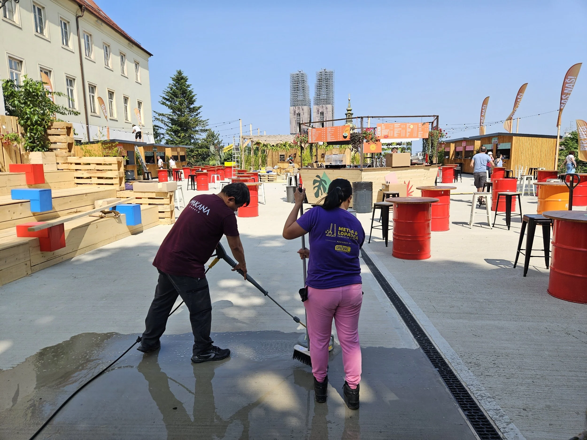 Two workers cleaning a sidewalk at an outdoor cafe or market area with red and black tables, stools, and wooden structures, under a blue sky, with construction in the background.
