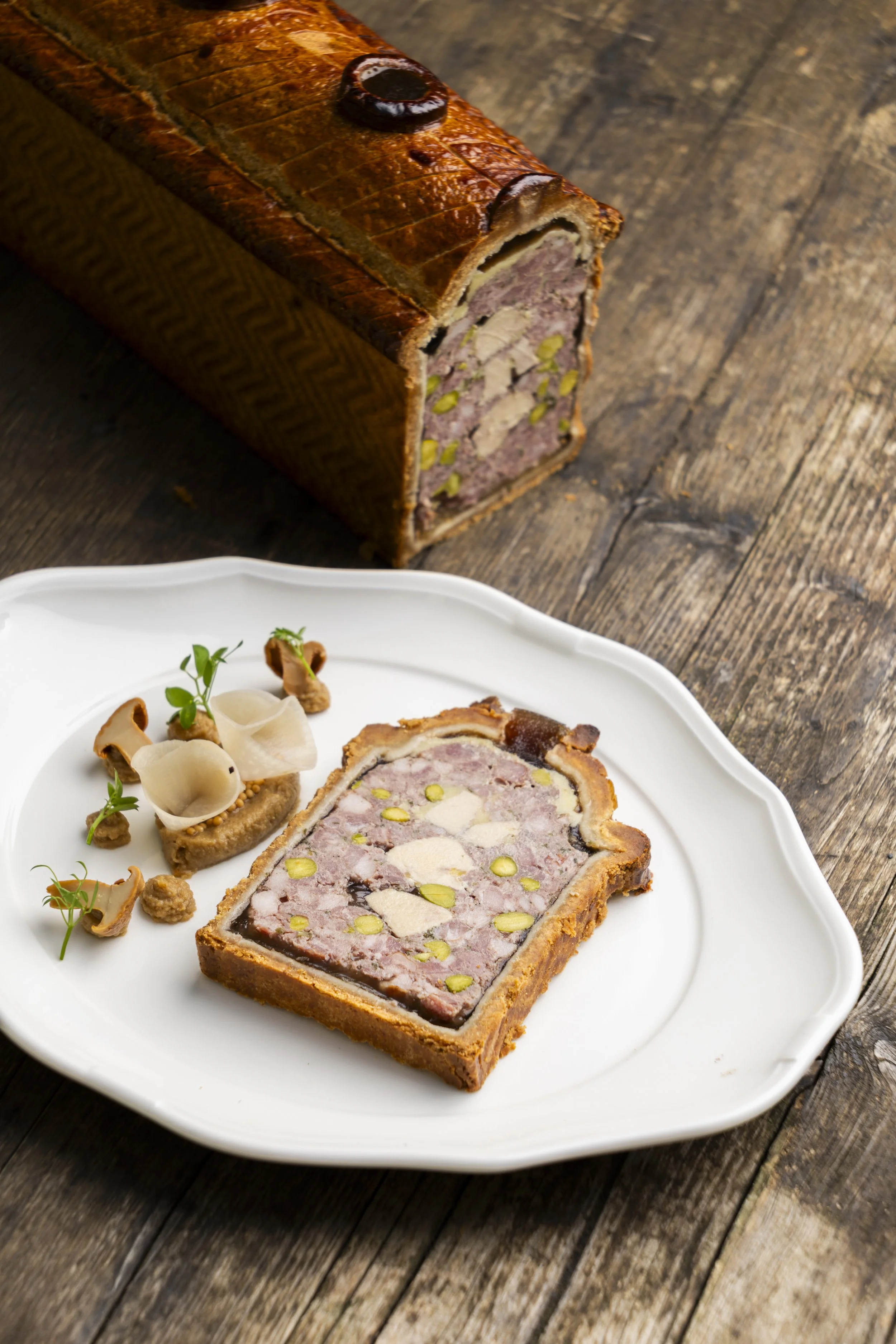 Pâté en croute présentée sur une assiette blanche avec des garnitures. La terrine est coupée pour révéler un intérieur contenant de la viande de porc, du foie gras et des pistaches, avec quelques tranches de champignons et des micro-pousses.