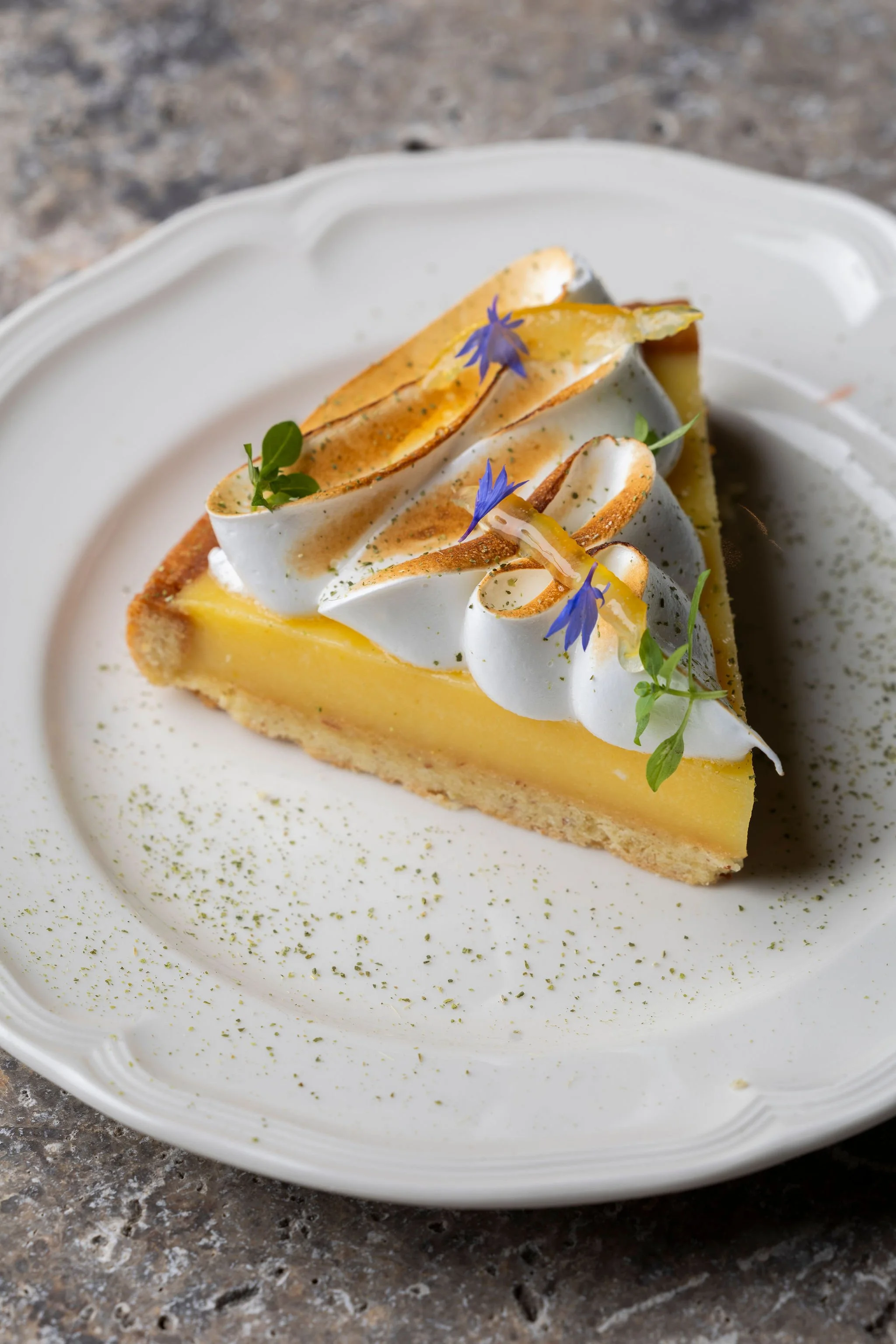 Une part de tarte au citron sur une assiette blanche, garnie de crème, de fleurs violettes et de feuilles vertes, avec une pincée de poudre verte