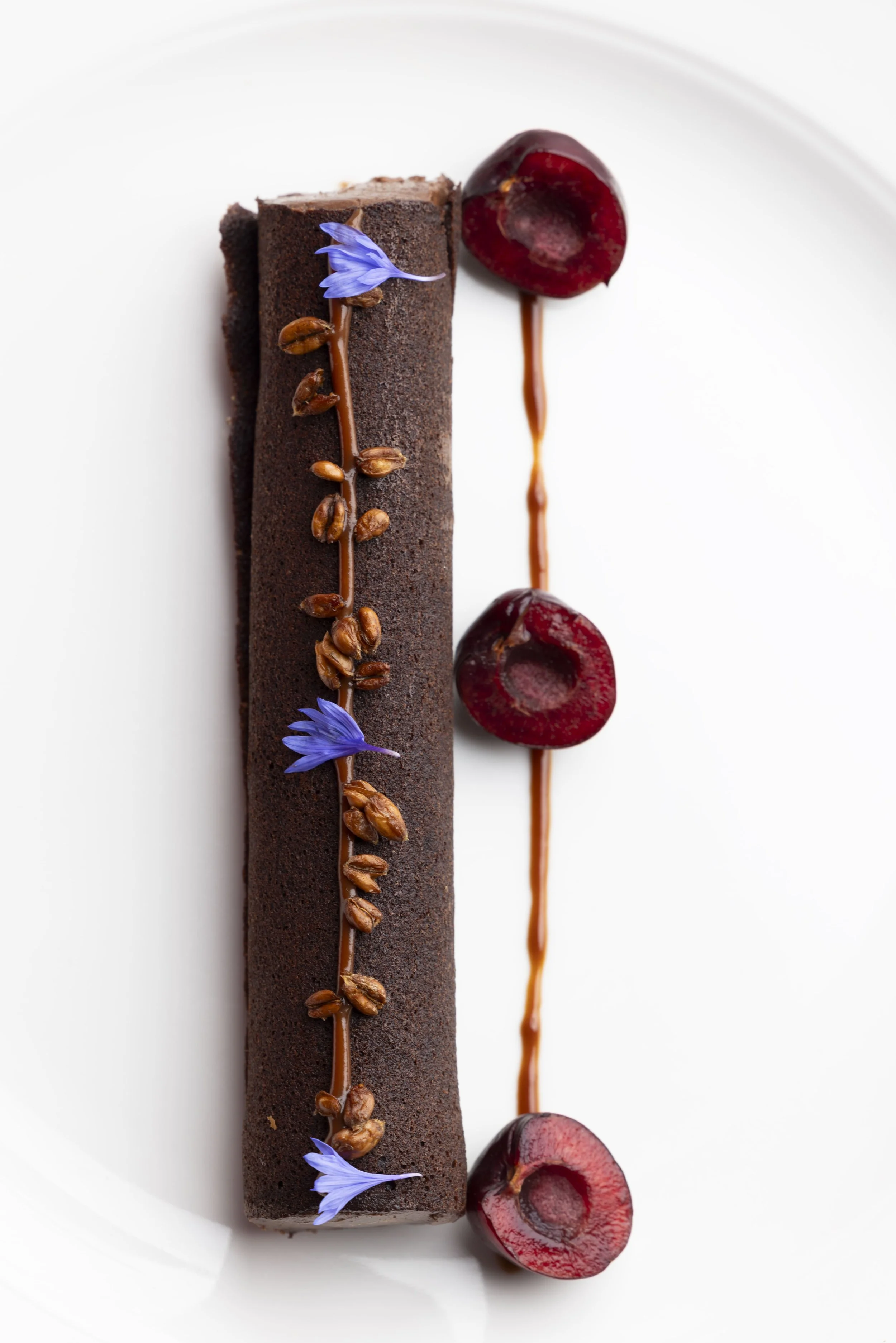 Un dessert au chocolat avec des cerises et des petites fleurs violettes sur une assiette blanche.