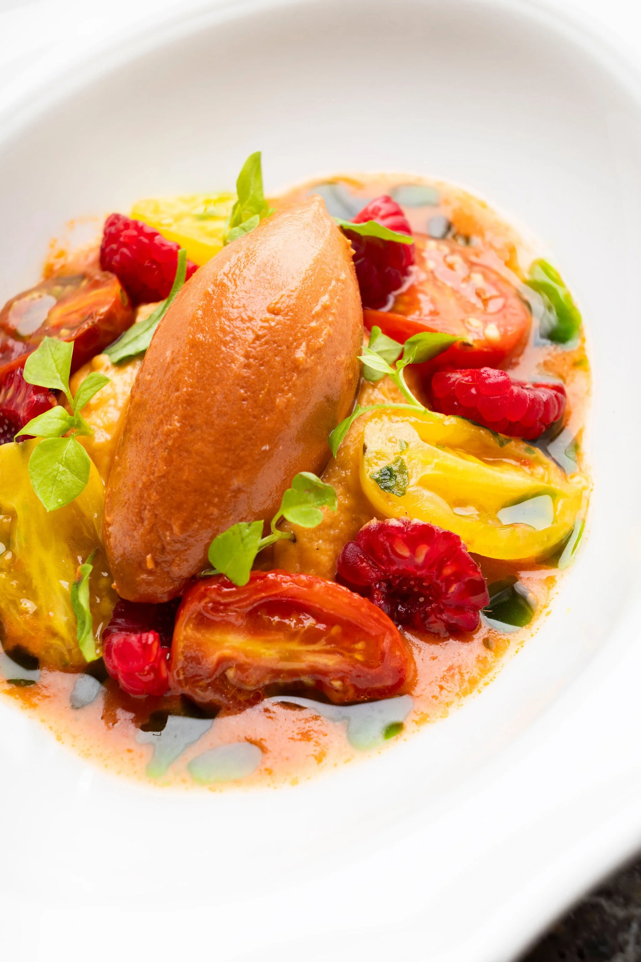 Assiette de soupe froide à la tomate, avec des morceaux de tomate, de fruits rouges, de basilic et une quenelle de crème, présentée dans une assiette blanche.
