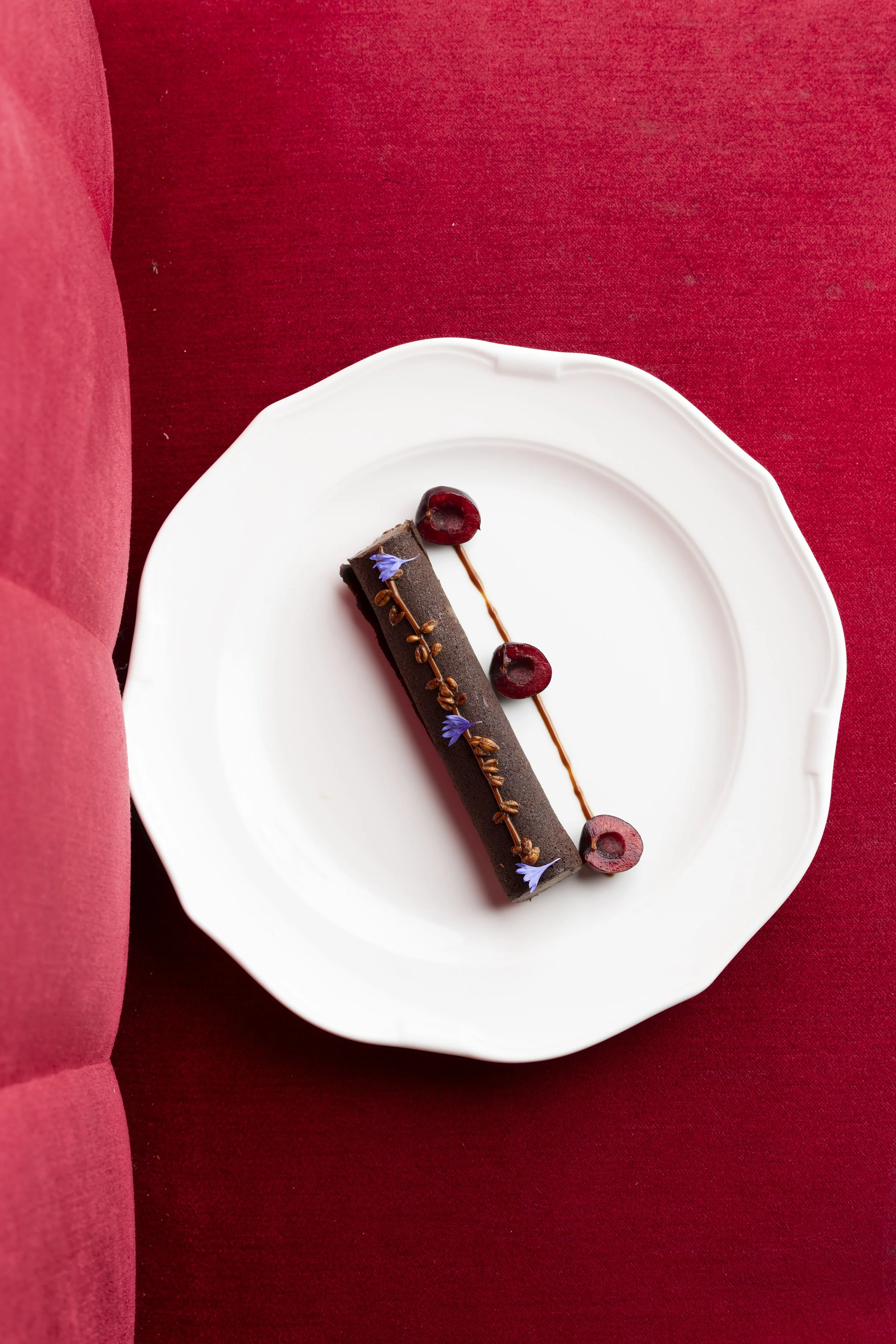 Un gâteau au chocolat rectangulaire décoré avec des myrtilles, des fleurs violettes et des petits éléments en chocolat, présenté sur une assiette blanche.