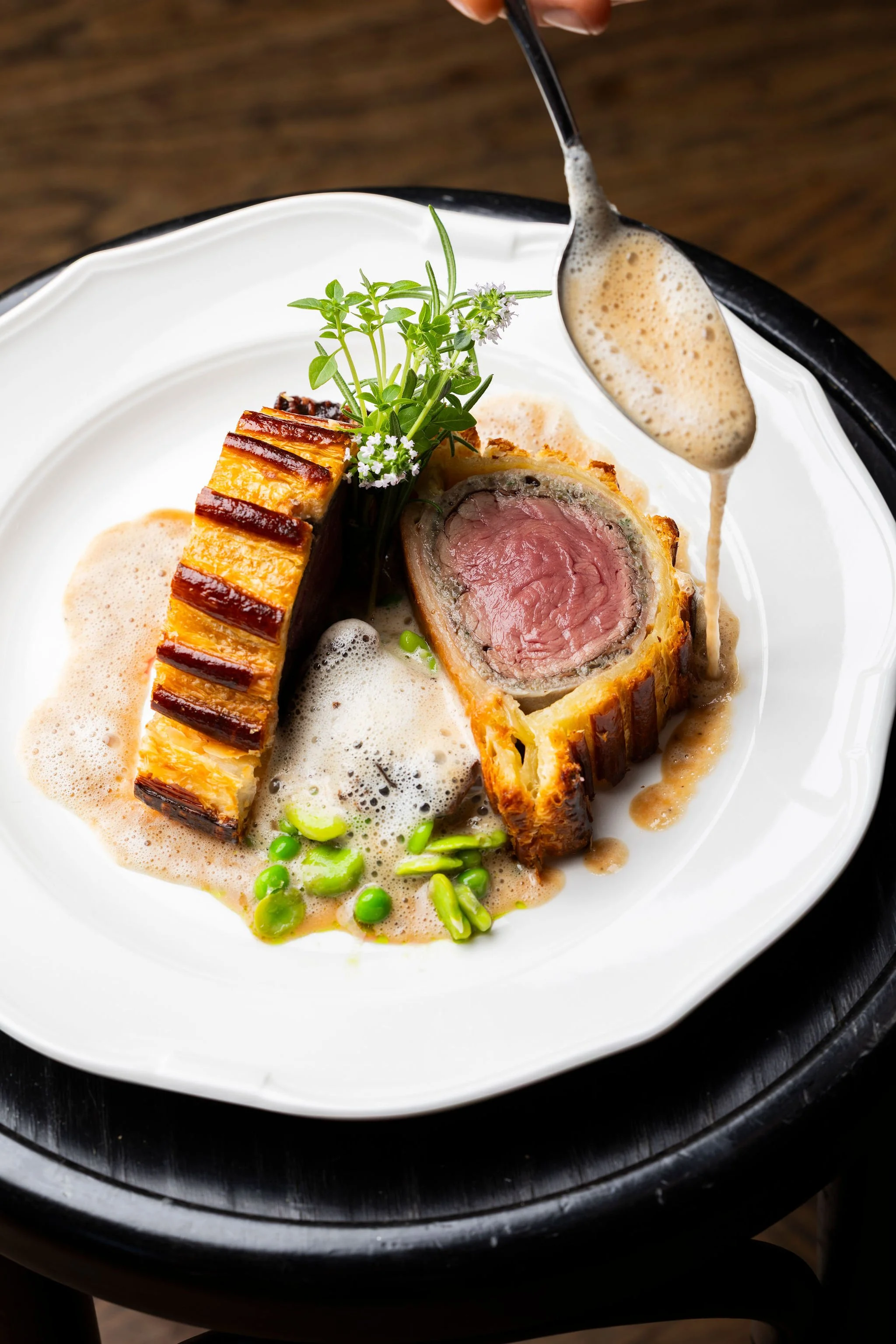 Plat gourmand présentant un boeuf wellington, accompagné de sauce, champignons et petits pois, décoré de fines herbes et fleurs