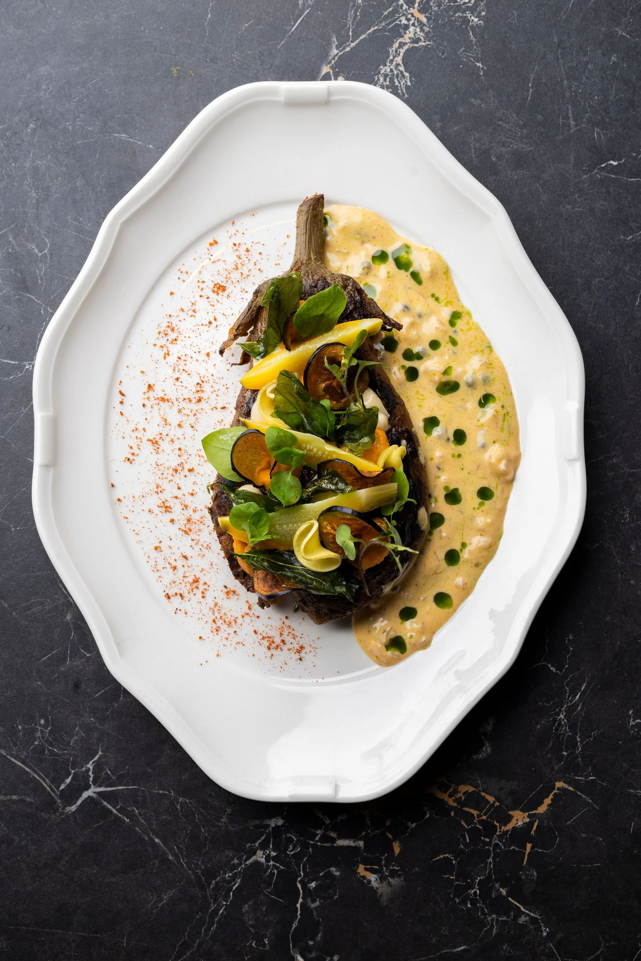 Plat de viande avec légumes et sauce crémeuse, décoré de fines herbes, présenté sur une assiette blanche avec une garniture rouge épicée, sur une surface en marbre noir.