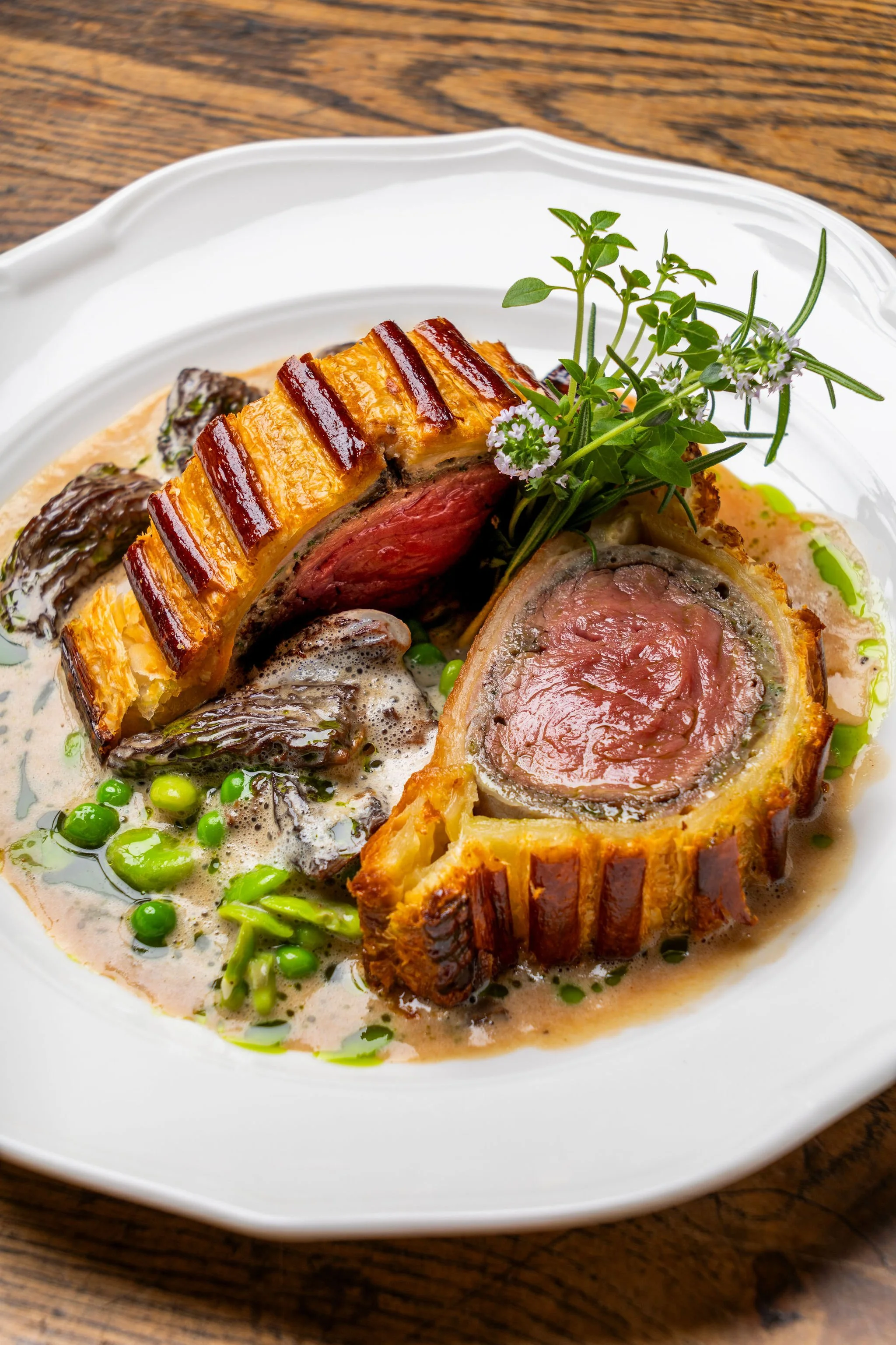 Plat gourmand présentant un boeuf wellington, accompagné de sauce, champignons et petits pois, décoré de fines herbes et fleurs