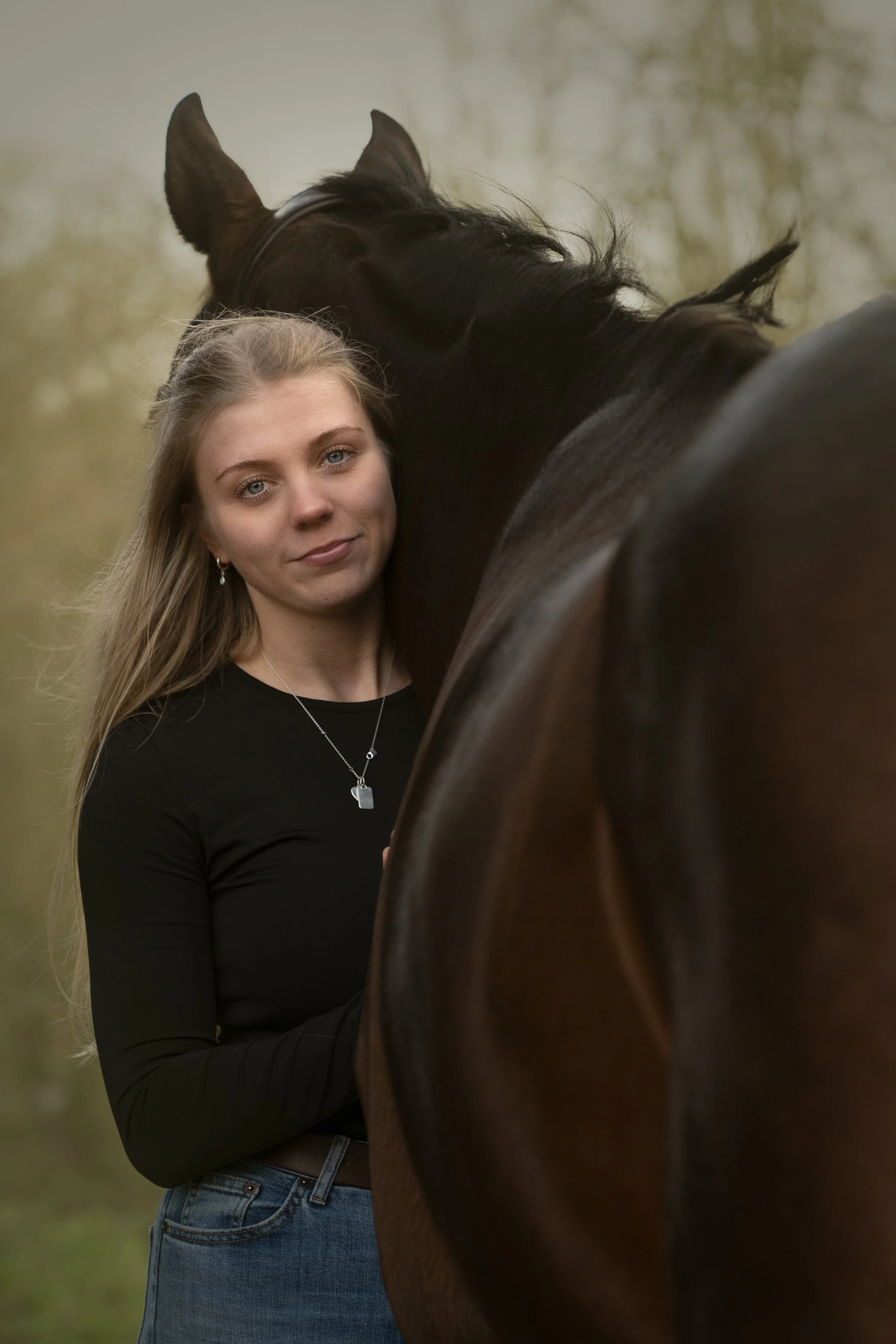 A young woman with long blonde hair, wearing a black long-sleeve top and jeans, standing close to a black horse outdoors with a blurred, natural background.