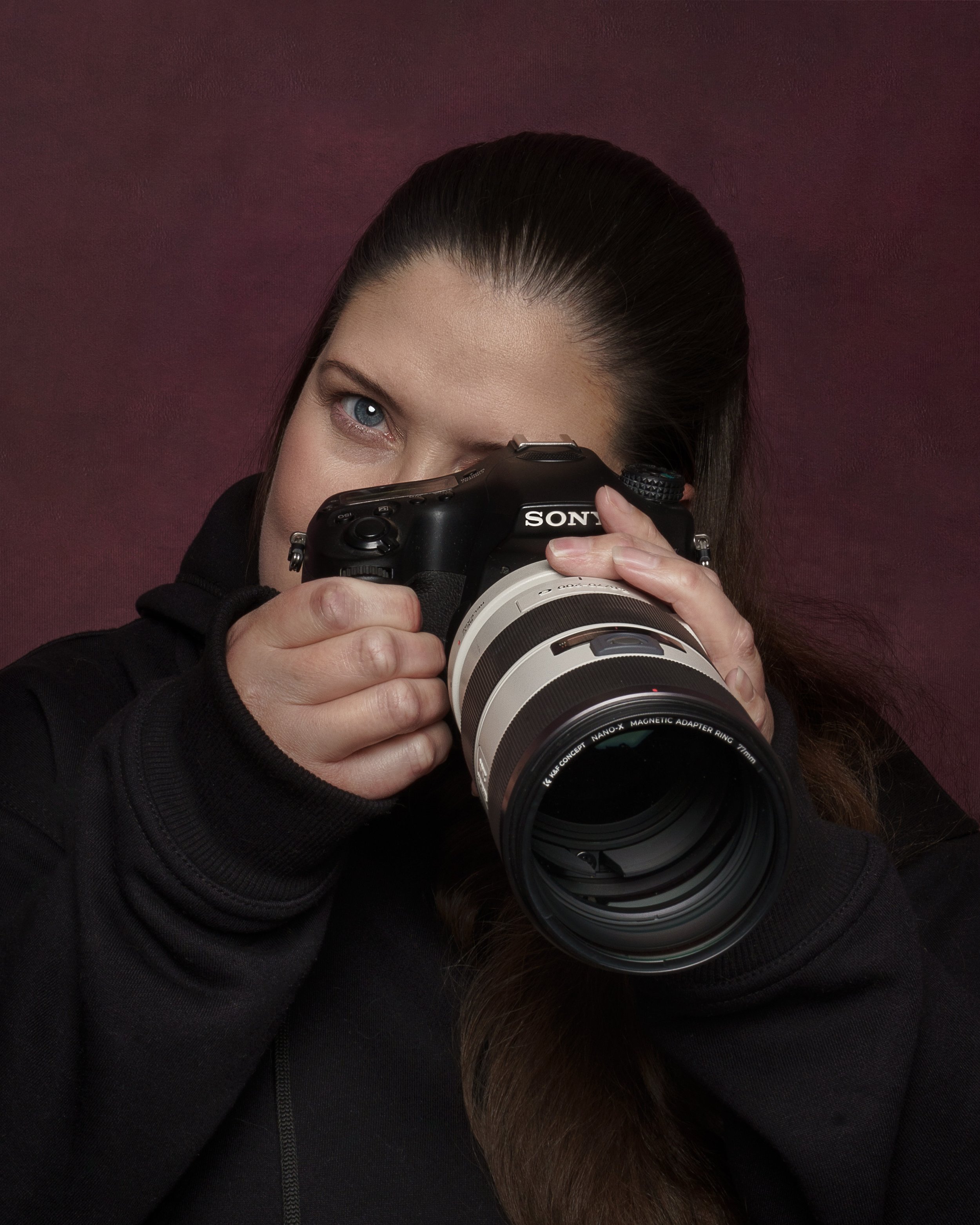 Woman with brown hair holding a Sony camera with a large lens, partially covering her face, against a dark maroon background.