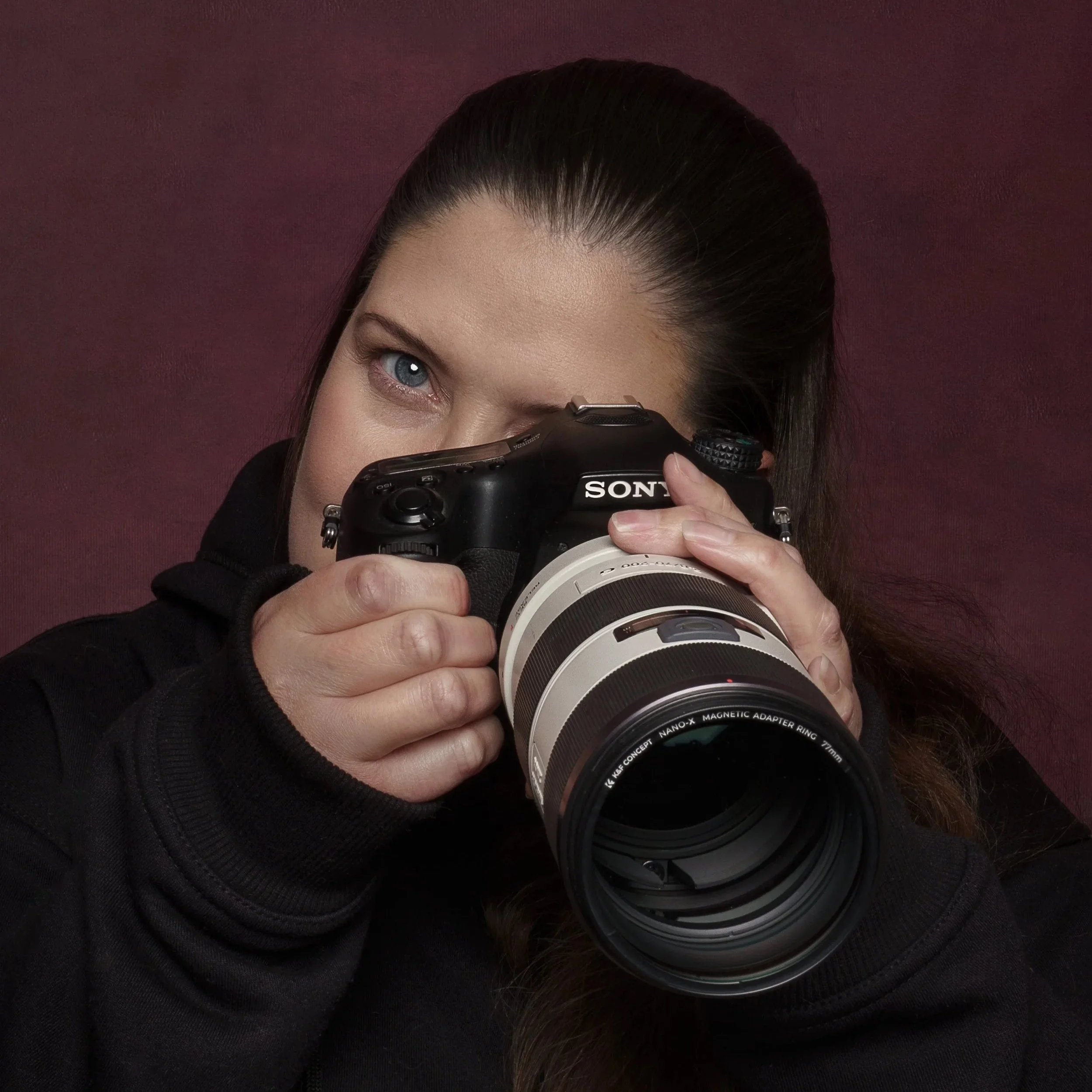 Woman with brown hair and blue eyes taking a photo with a professional camera against a dark red background.