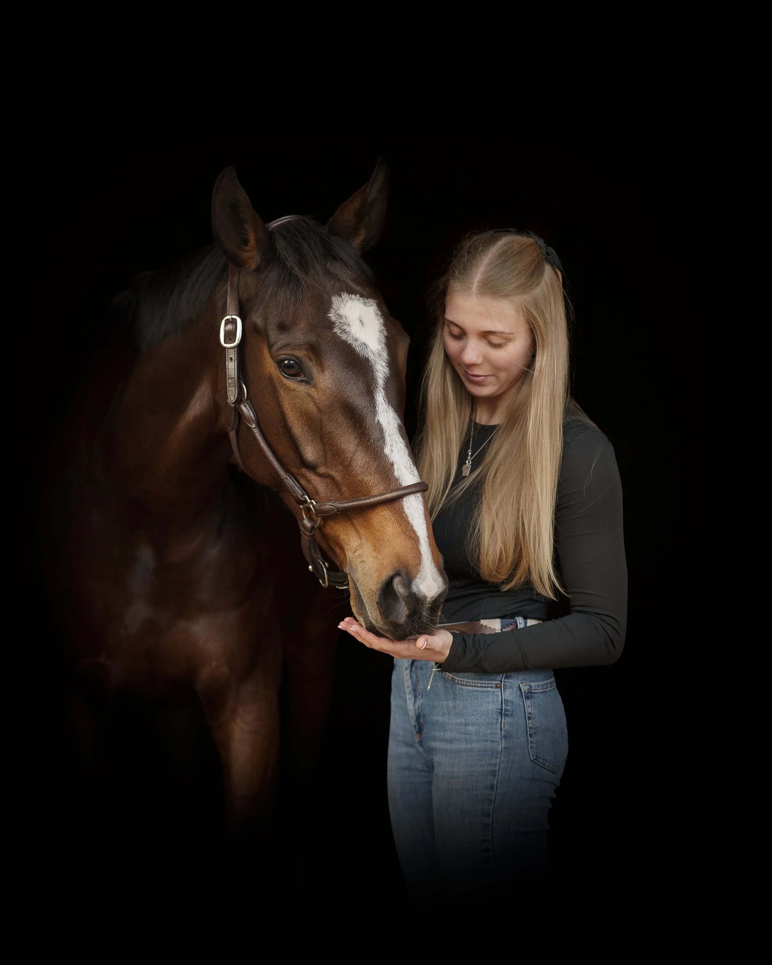 A young woman in a black long-sleeve shirt and jeans gently holds a brown horse's nose, both sharing a peaceful moment against a black background.