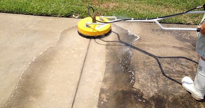 A person using a yellow pressure washer to clean a concrete driveway, with water visible on the surface and grass in the background.