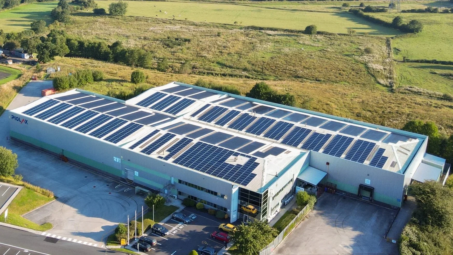 A large industrial building with solar panels on the roof, surrounded by parking lots and green landscape.