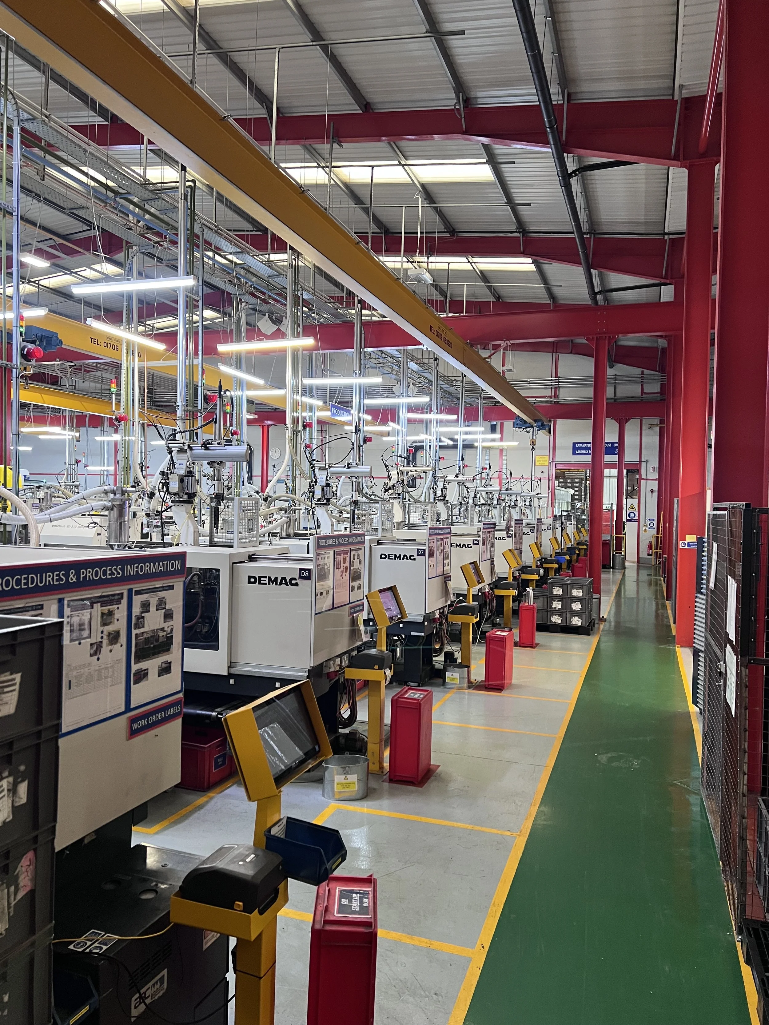 Factory or industrial workspace with multiple automated machines and control panels, yellow safety barriers, red emergency fire extinguishers, and overhead cranes in a large, well-lit environment.