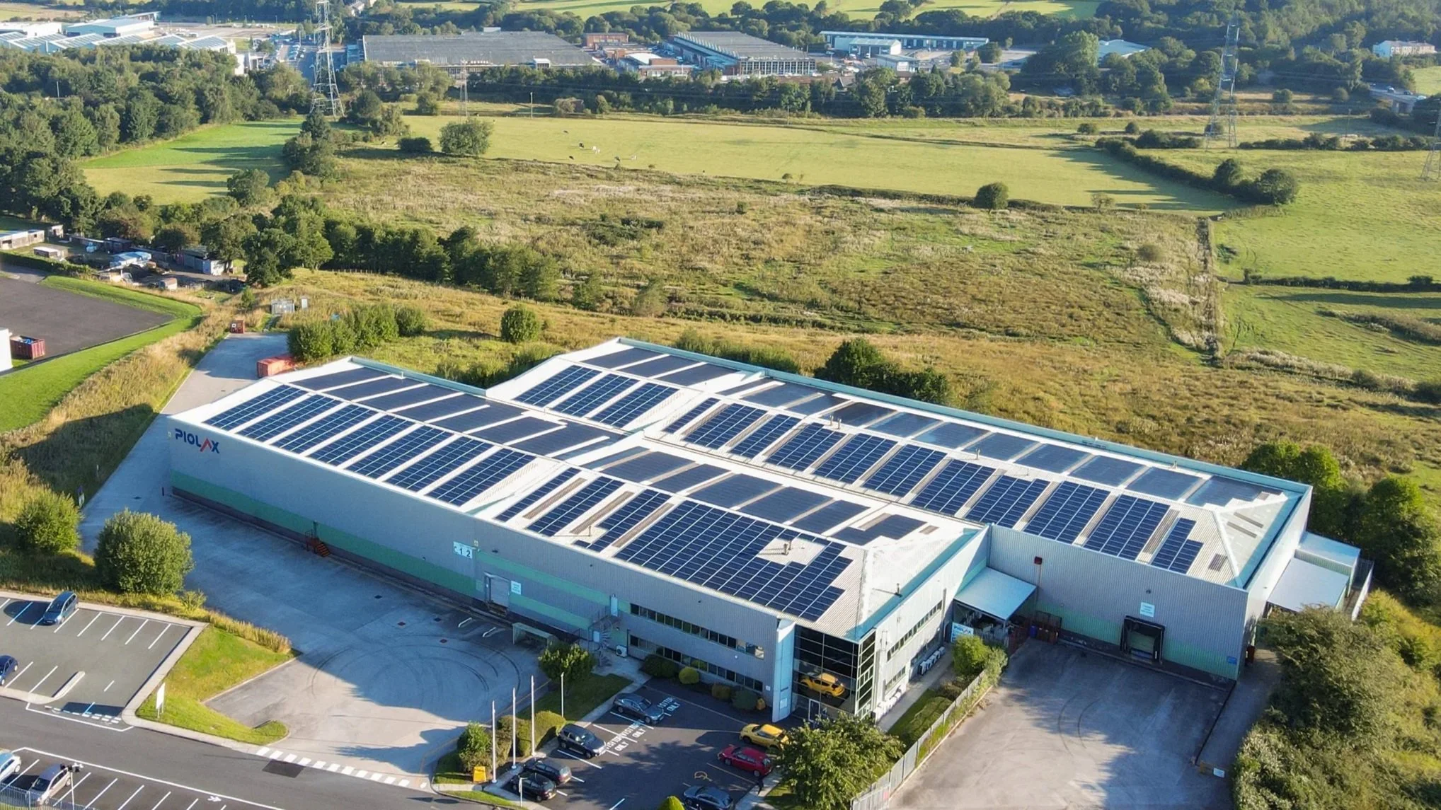 Aerial view of a large industrial building with solar panels on the roof, surrounded by parking lot and green fields.