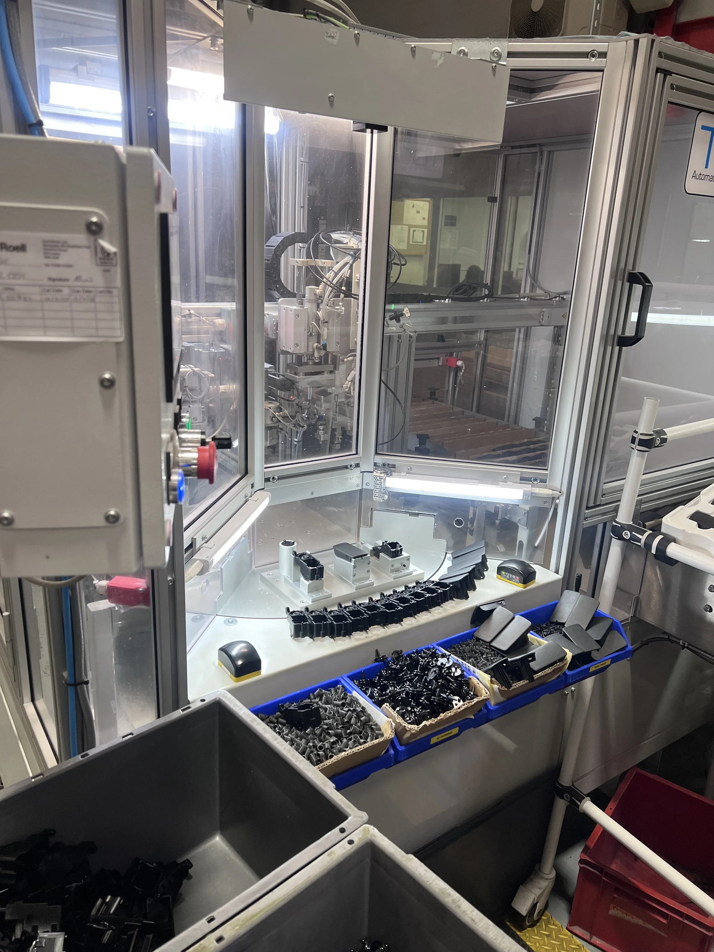 A manufacturing machine with multiple containers of black plastic components and metal parts inside a factory or workshop.