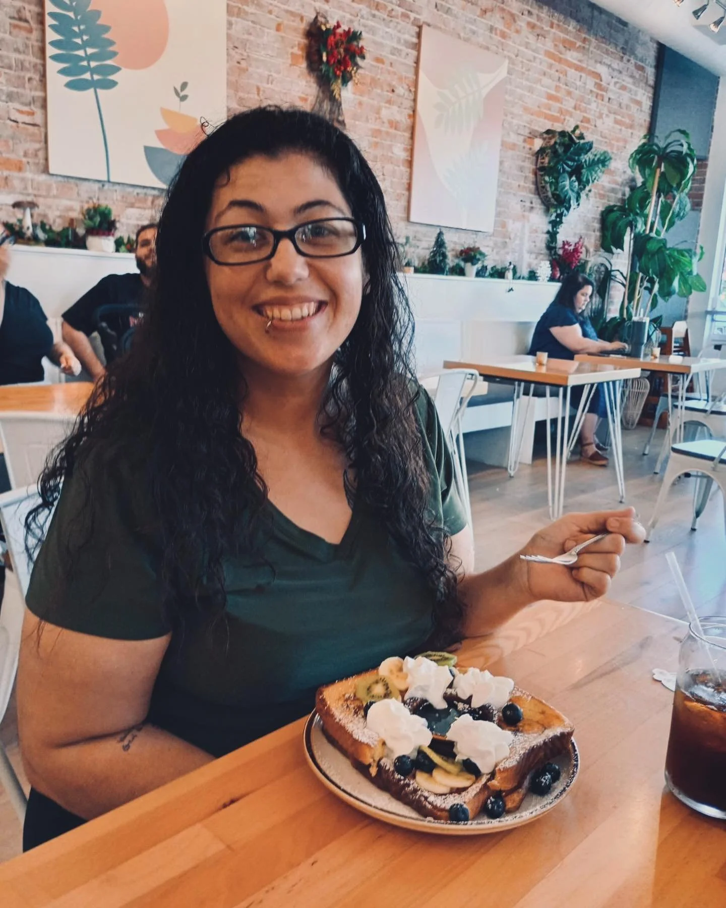 A woman with glasses smiling, sitting at a table with a plate of French toast topped with whipped cream, fruit, and powdered sugar, and holding a fork, in a cafe with brick walls and artwork.