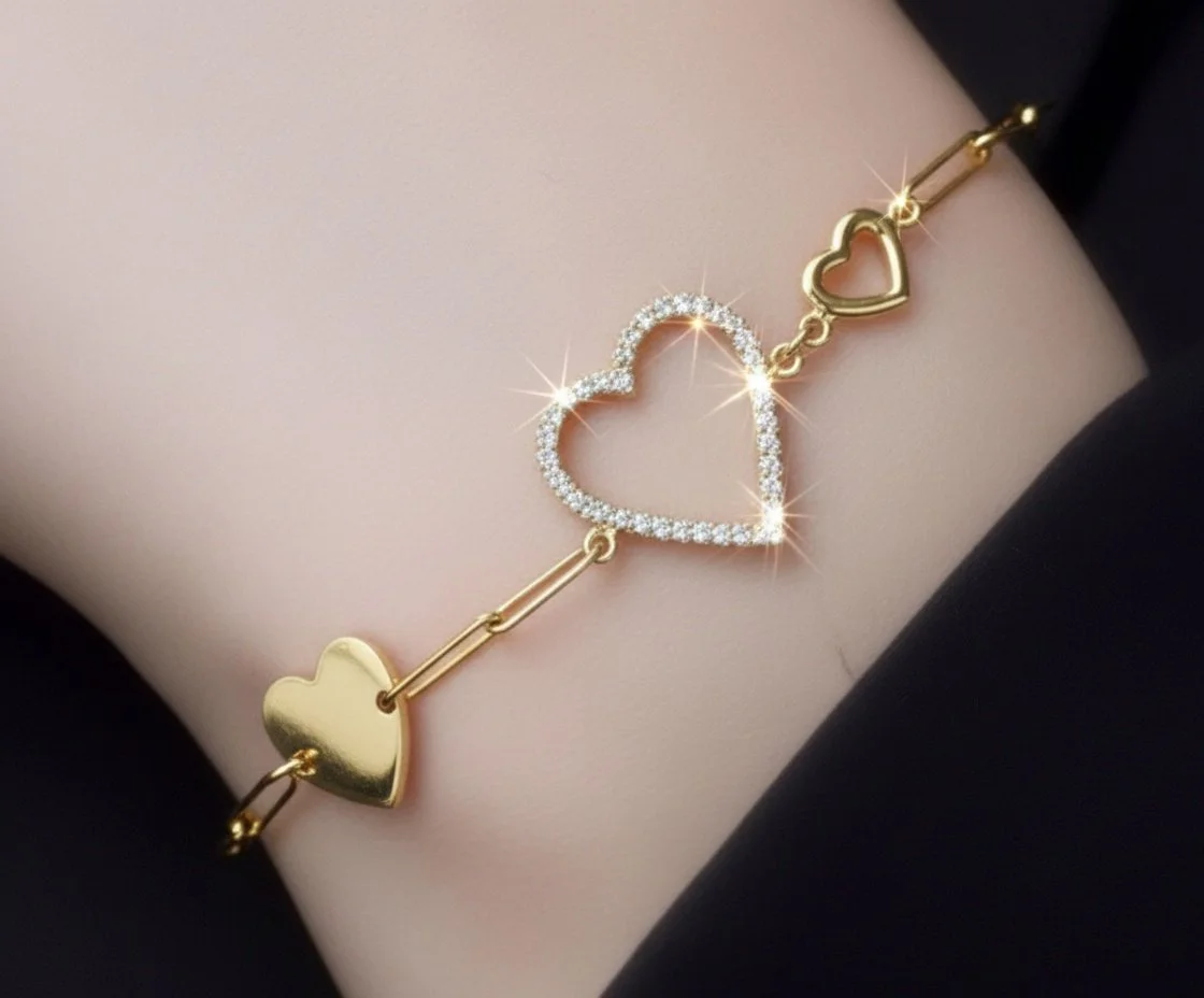 A gold bracelet with outlined hearts, one of which is encrusted with small gemstones, worn on a person's wrist.