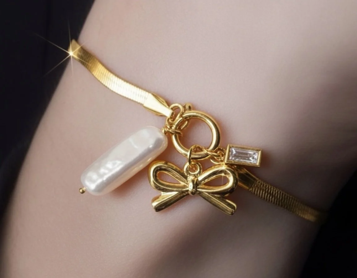 A gold bracelet with charms, including a white pearl-like bead, a gold bow, and a small rectangular clear gemstone, worn around a person's wrist.