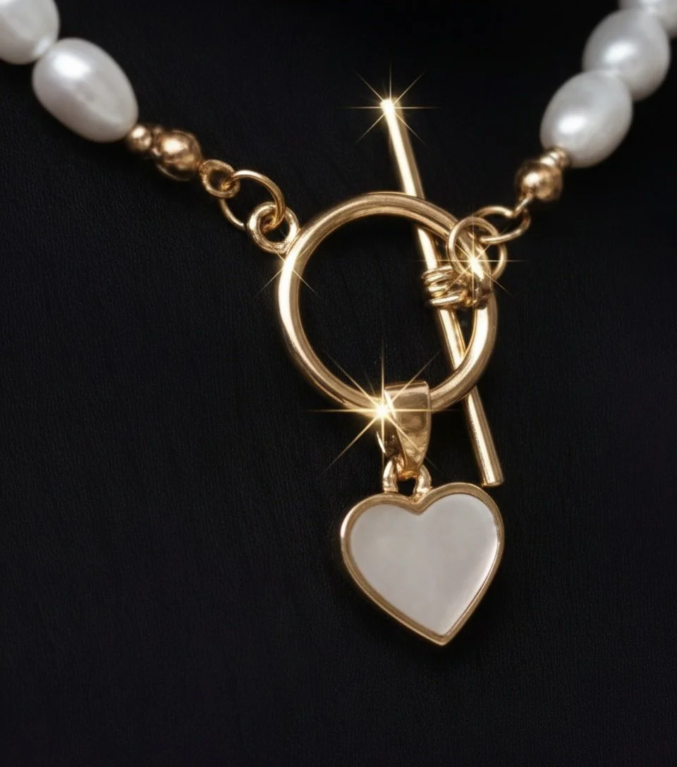 Close-up of a pearl and gold necklace with a heart-shaped pendant and a toggle clasp, against a black background with bright reflections.