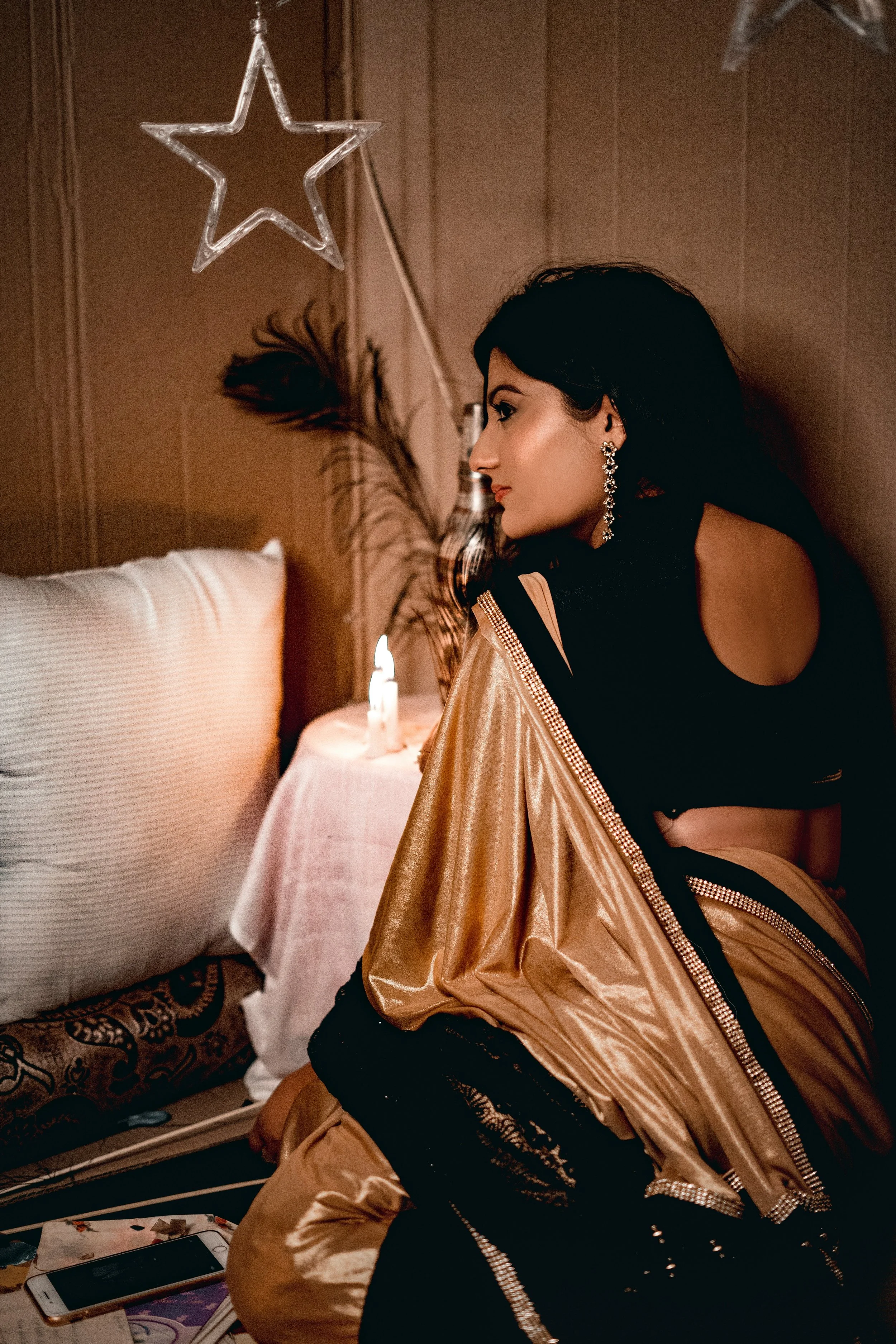 A woman with dark hair and earrings sitting on a bed, dressed in a shiny gold jacket and black lace pants, looking to the side in a dimly lit room with a star-shaped light fixture and candles.