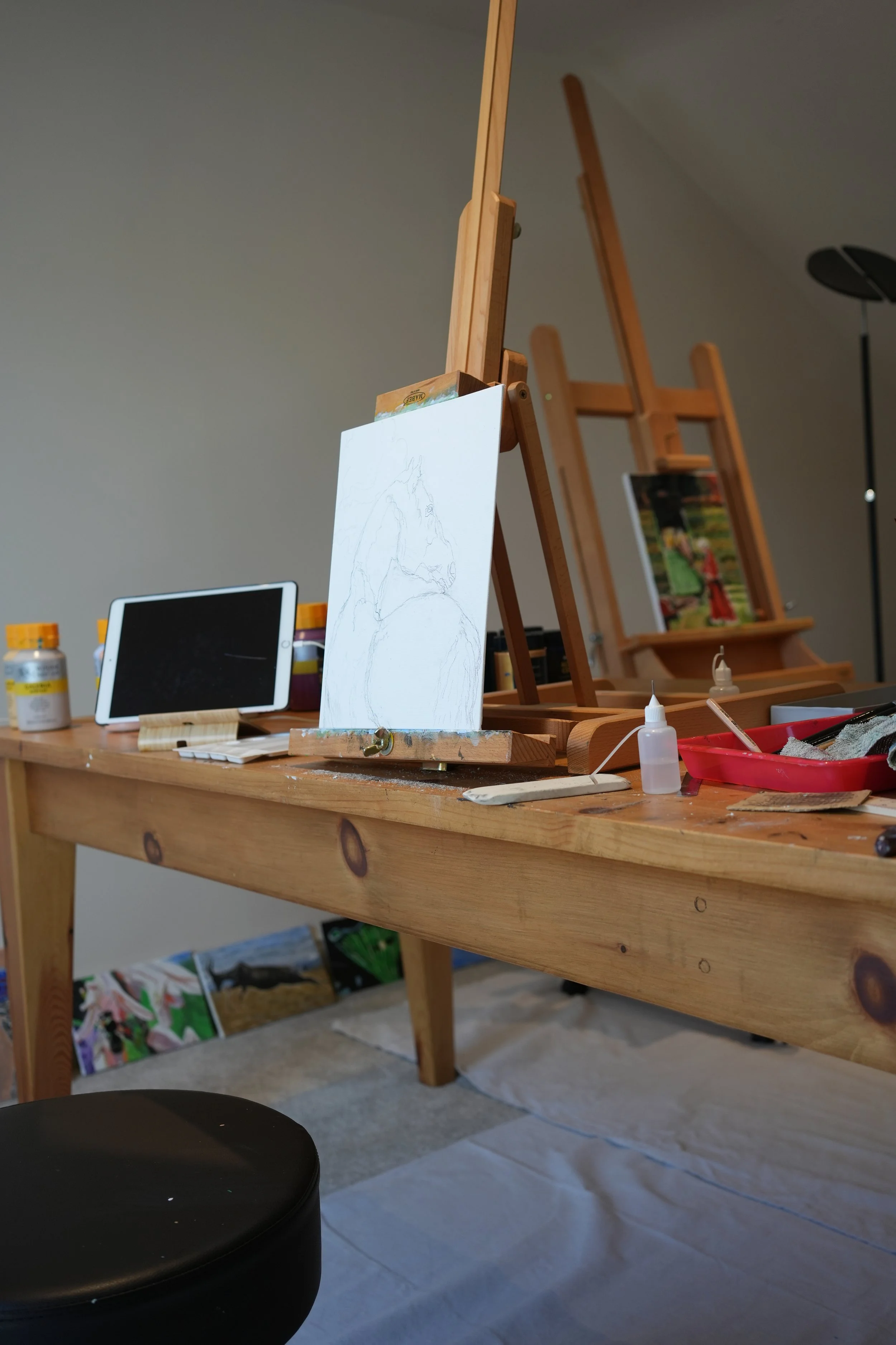 Art studio with a wooden table holding an easel with a canvas, art supplies, and a tablet, with another easel and artwork in the background.