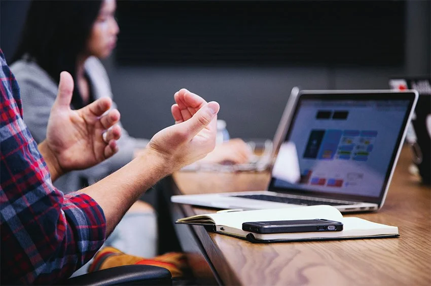 Person gesturing with hands in a business meeting with a laptop, notebook, and phone on the table.