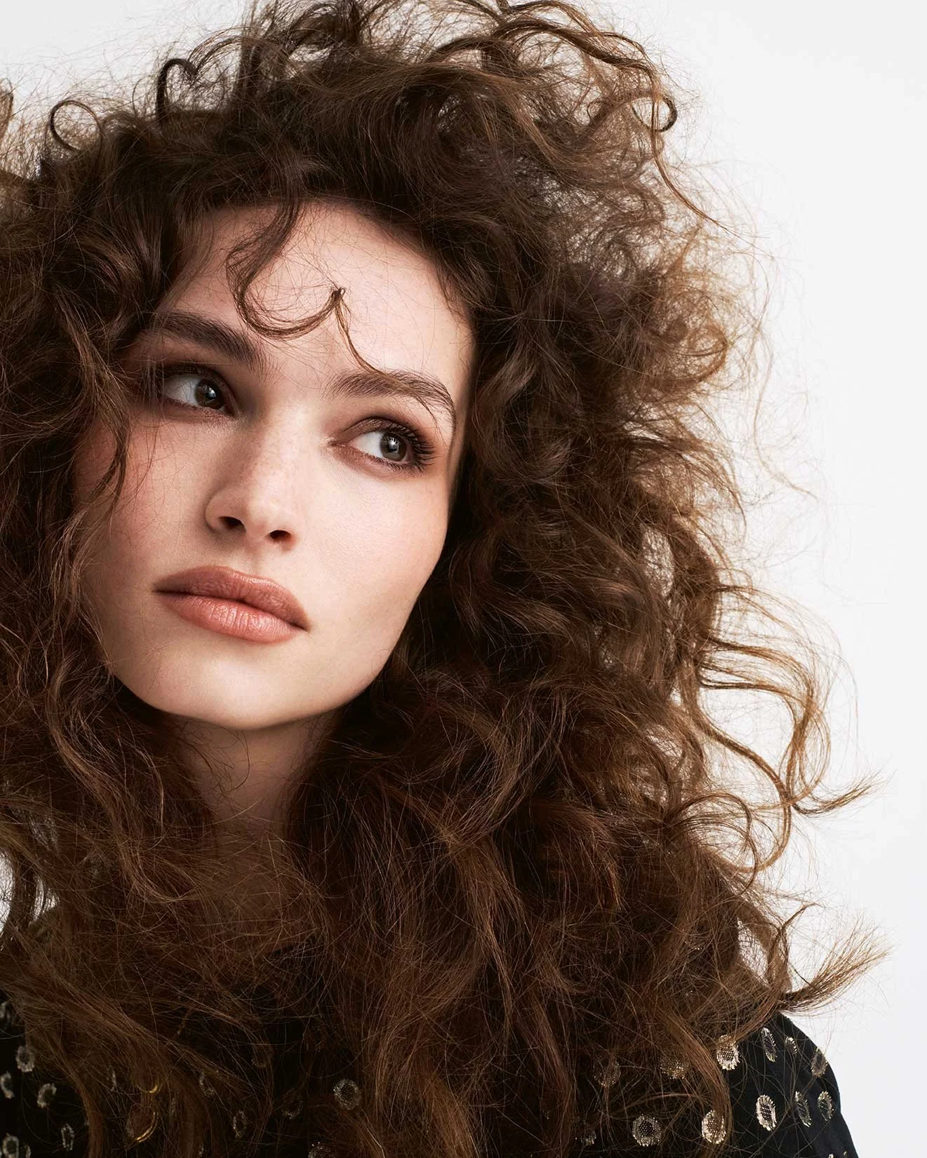 Porträt einer jungen Frau mit langen, lockigen braunen Haaren, blasser Haut, dezenten Make-up und einem schwarzen Oberteil mit goldenen Pailletten.