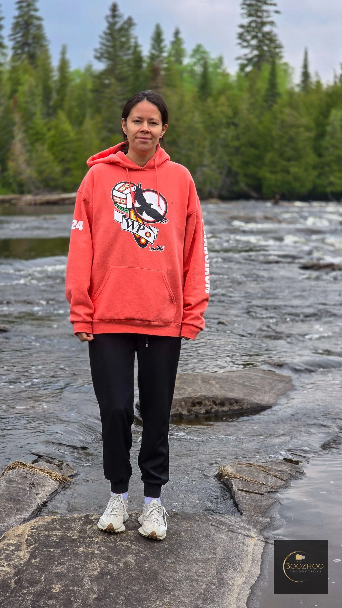 A woman standing on rocks in a river with trees and cloudy sky in the background, wearing a red hoodie with a black logo, black pants, and white sneakers.