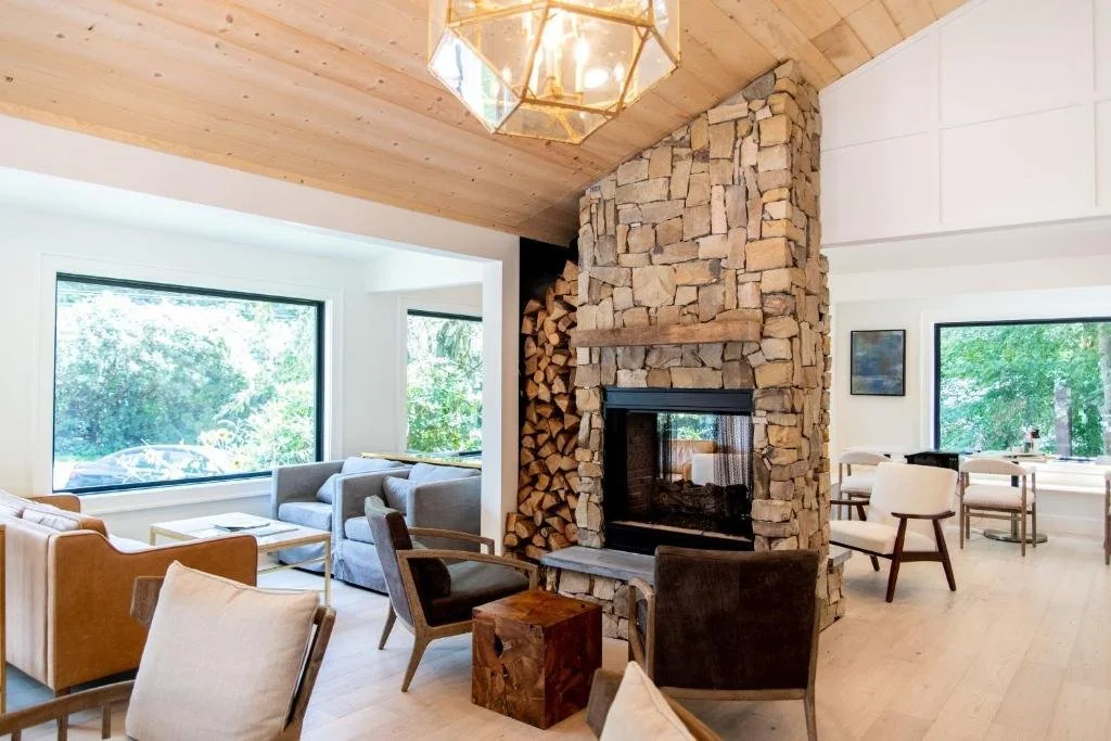 Living room with a stone fireplace, wooden ceiling, large windows showing greenery outside, and various chairs and sofas.