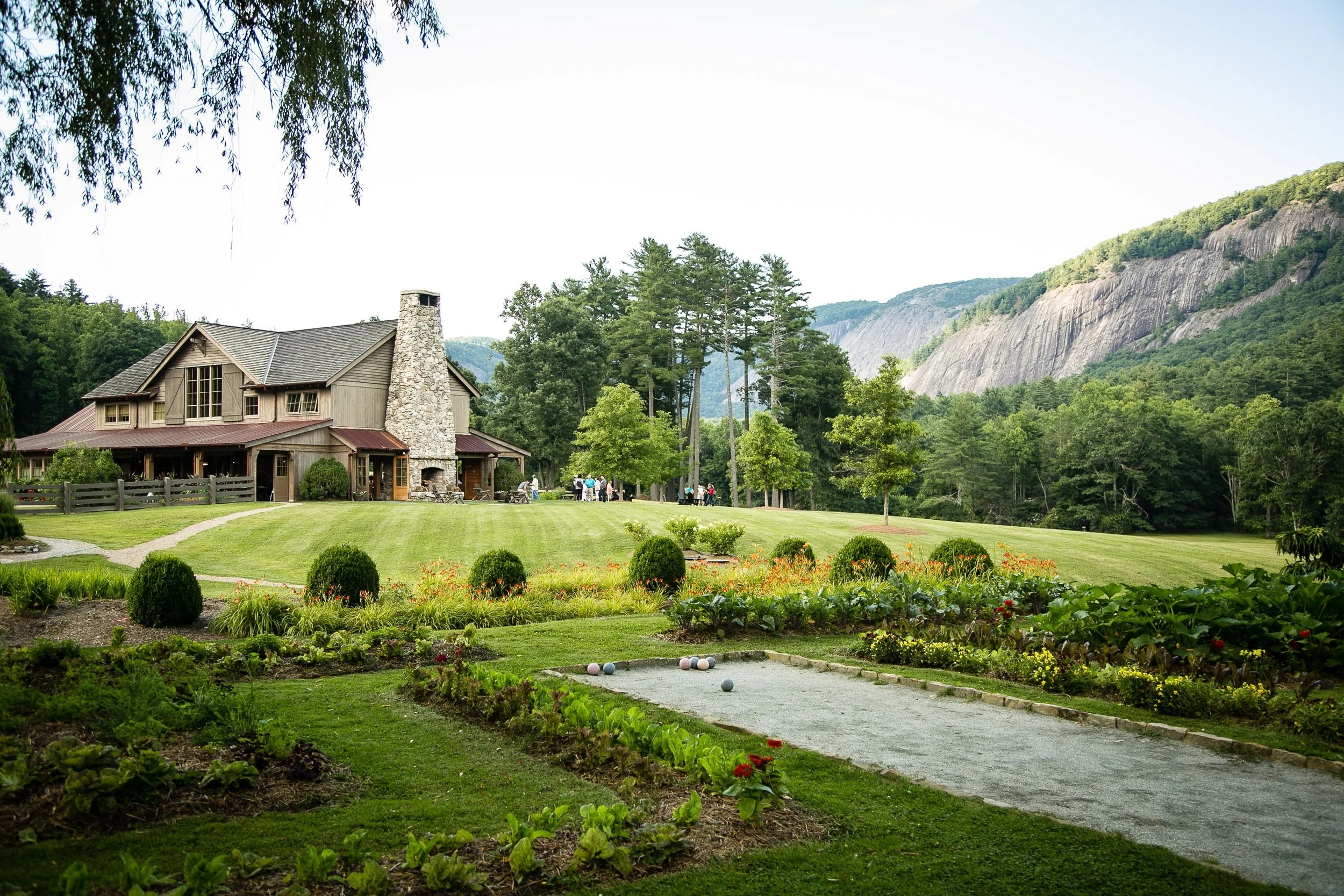 A large stone house with a chimney and expansive lawn, surrounded by trees and mountains, with a bocce ball court and planted flowers in the foreground.